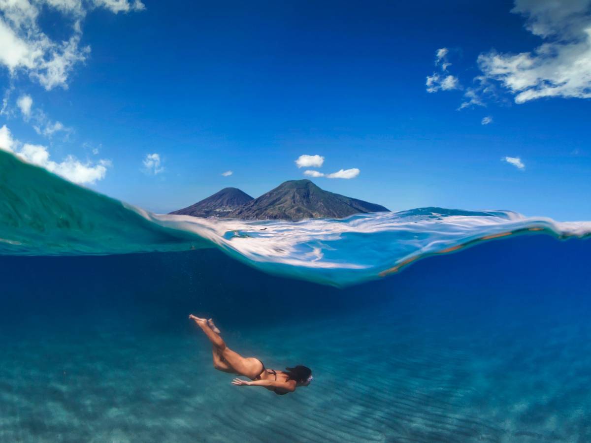 Eine Frau taucht im Meer, über ihr ist eine Insel mit zwei Vulkanen zu sehen.