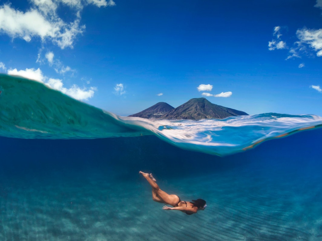 Eine Frau taucht im Meer, über ihr ist eine Insel mit zwei Vulkanen zu sehen.