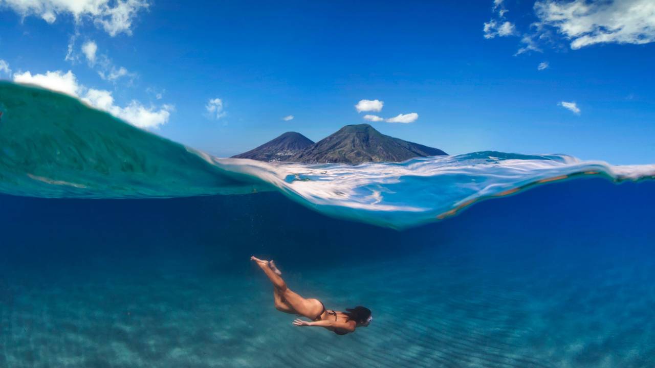 Eine Frau taucht im Meer, über ihr ist eine Insel mit zwei Vulkanen zu sehen.