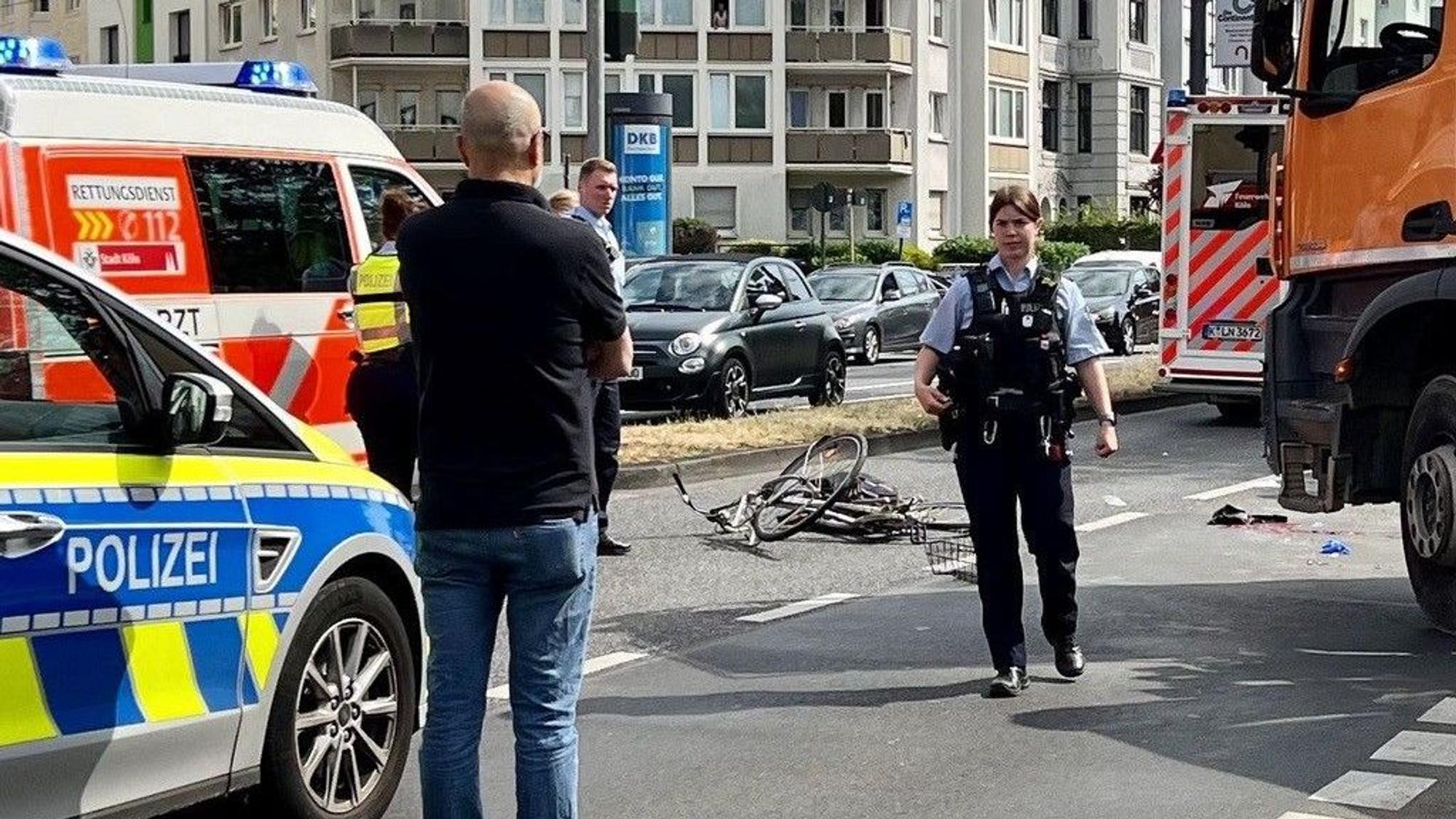 Ein zerbeultes Fahrrad liegt auf der Fahrbahn, dort stehen auch Einsatzkräfte und Einsatzfahrzeuge von Polizei und Rettungsdienst.