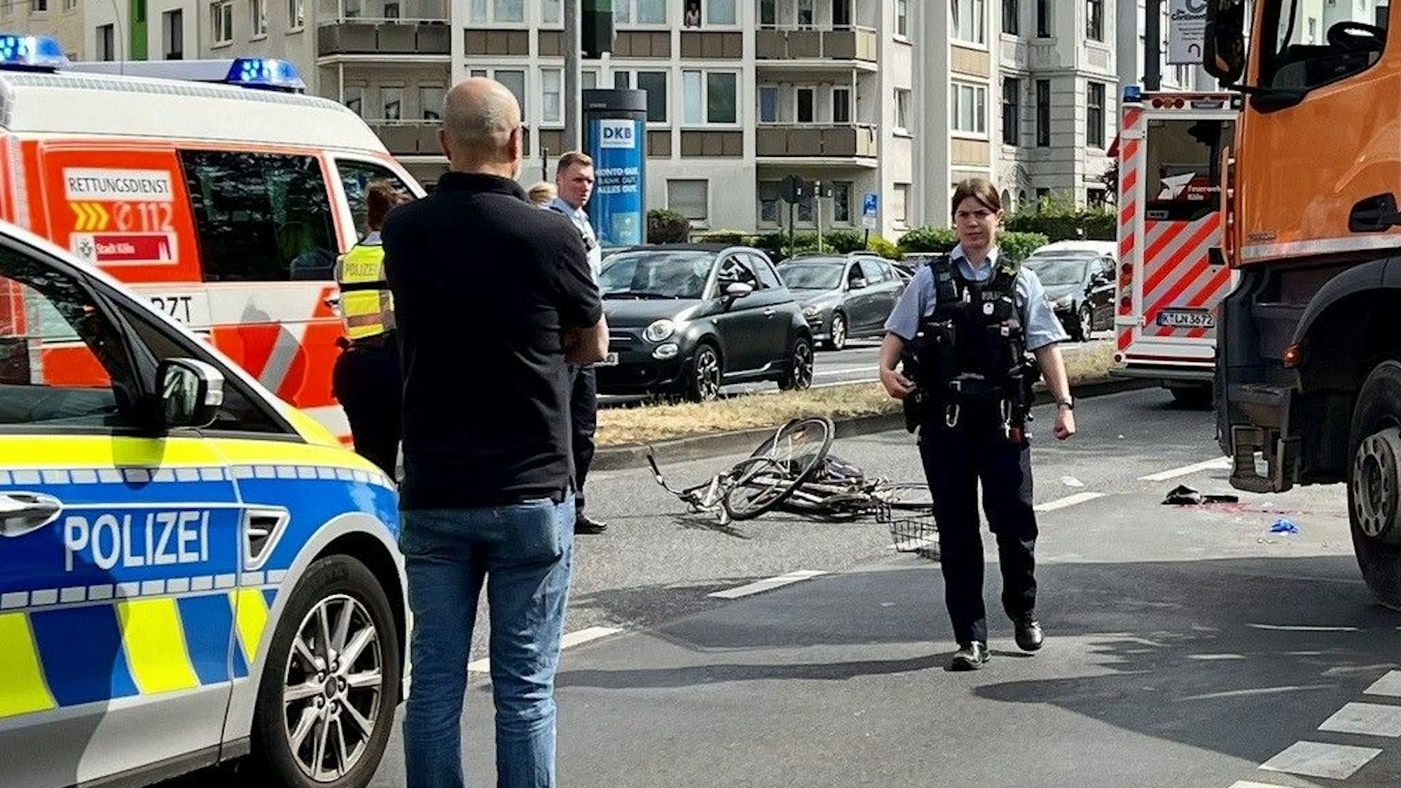 Ein zerbeultes Fahrrad liegt auf der Fahrbahn, dort stehen auch Einsatzkräfte und Einsatzfahrzeuge von Polizei und Rettungsdienst.
