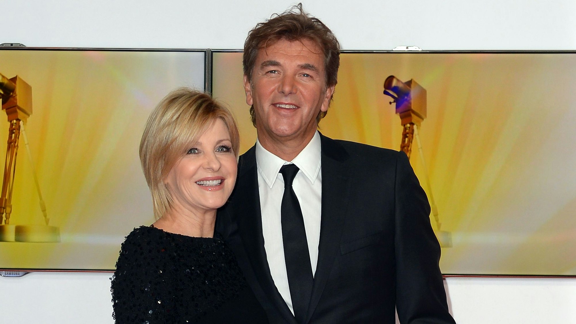 Carmen Nebel und Norbert Endlich posieren lächelnd in Gala-Kleidung auf dem roten Teppich der Goldenen Kamera