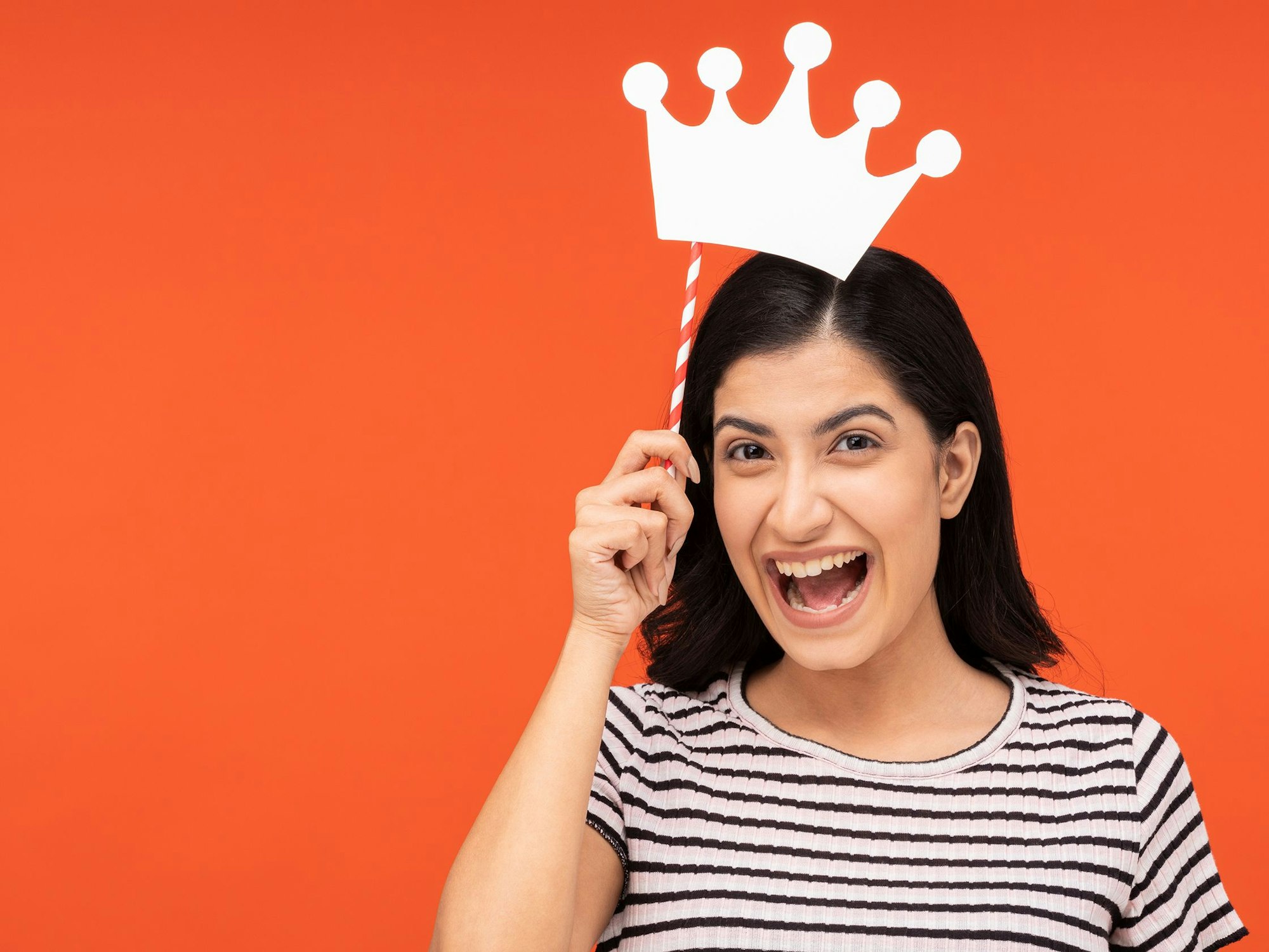 Eine lachende Frau hält sich eine stilisierte Krone am Stiel über den Kopf.