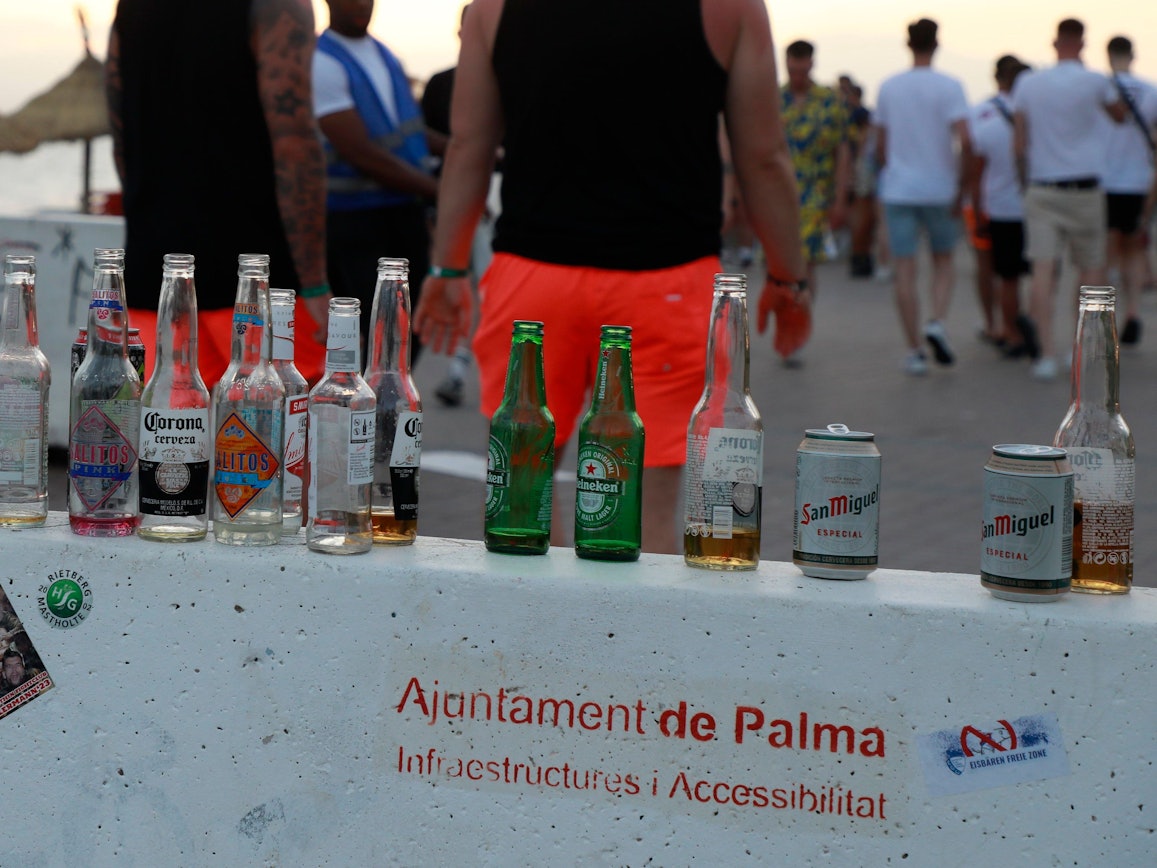 Menschen haben sich am Abend an der Promenade am Strand von Arenal versammelt. Auf einer Mauer stehen mehrere leere Flachen alkoholischer Getränke.