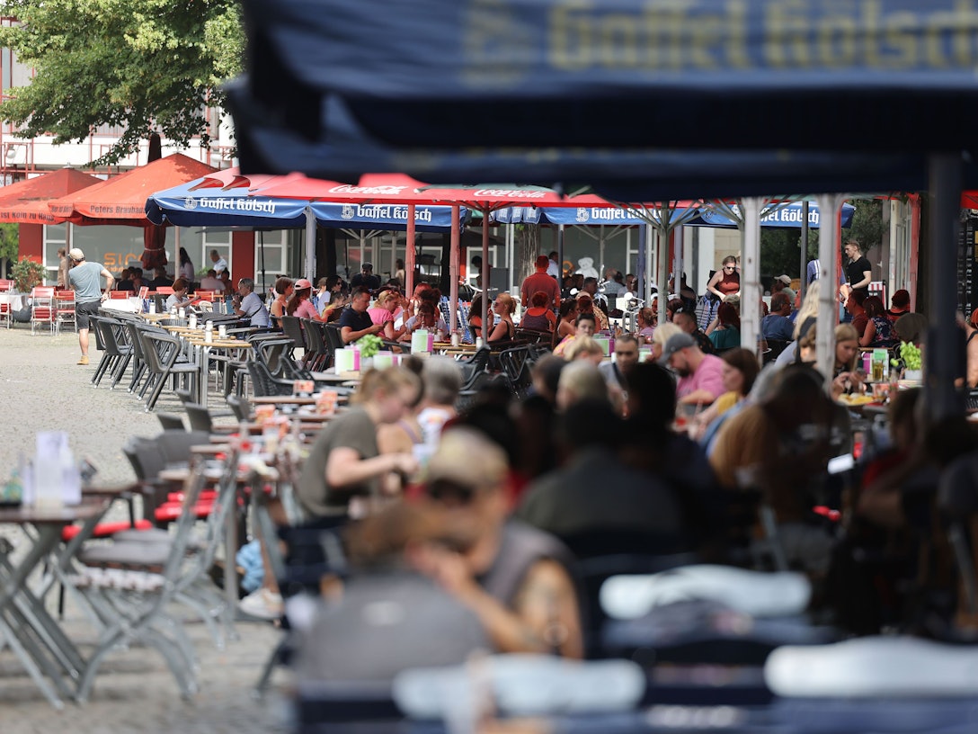 Menschen sitzen in Lokalen im Außenbereich unter Sonnenschirmen. Ab diesem Jahr gelten neue, verbindliche Regeln für die Außengastronomie. Lokale in der Altstadt und im Bereich der Deutzer Messe müssen die Vorgaben ab dem 1. Oktober umsetzen.
