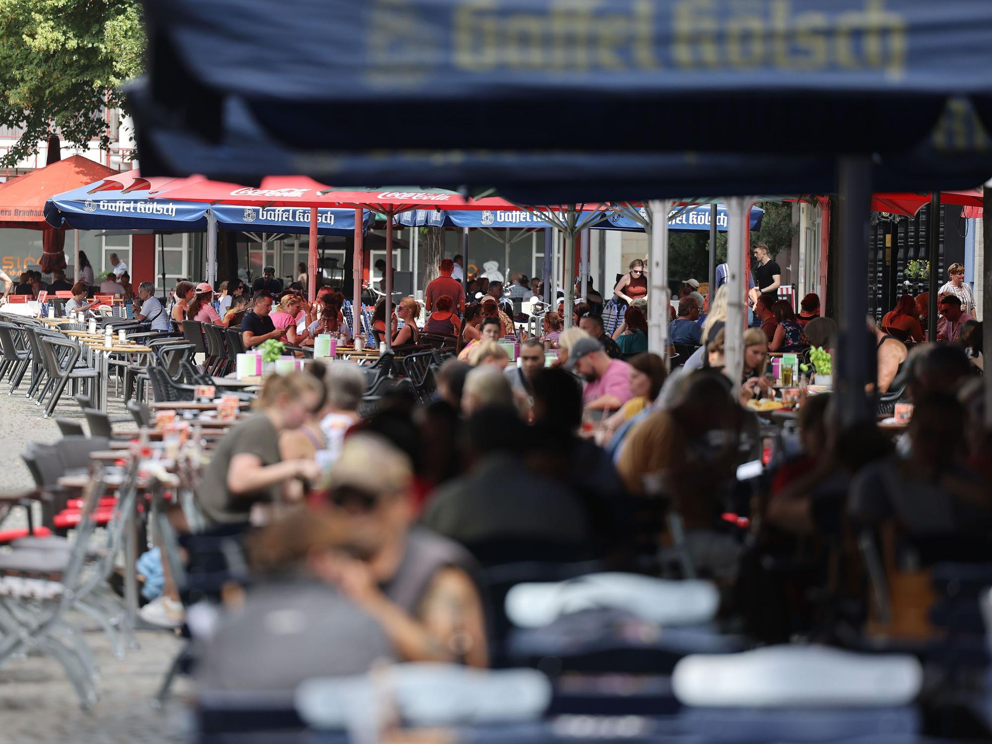 Menschen sitzen in Lokalen im Außenbereich unter Sonnenschirmen. Ab diesem Jahr gelten neue, verbindliche Regeln für die Außengastronomie. Lokale in der Altstadt und im Bereich der Deutzer Messe müssen die Vorgaben ab dem 1. Oktober umsetzen.