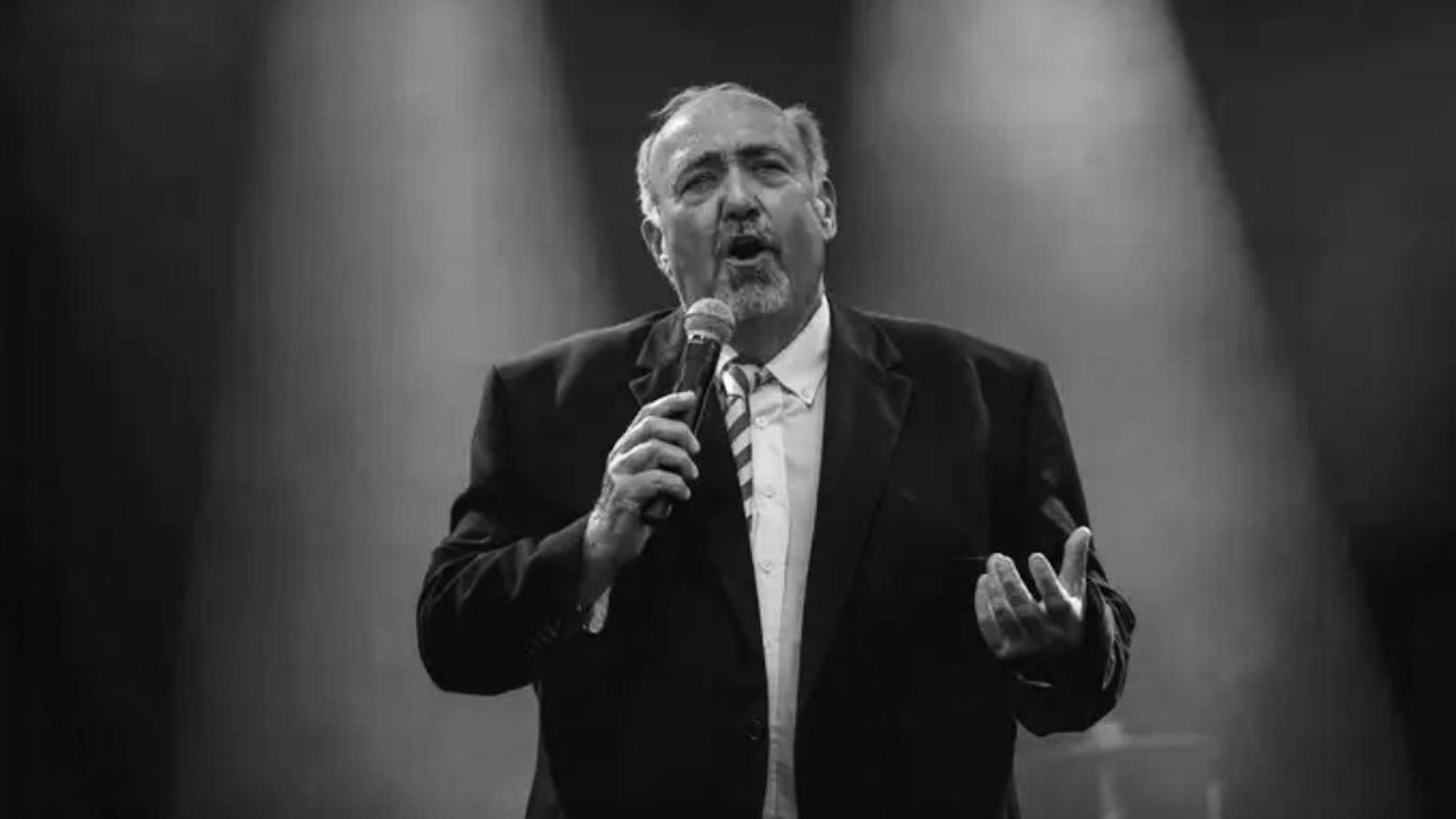 Der verstorbene Bärchen Sester steht mit Mikrofon auf einer Bühne.