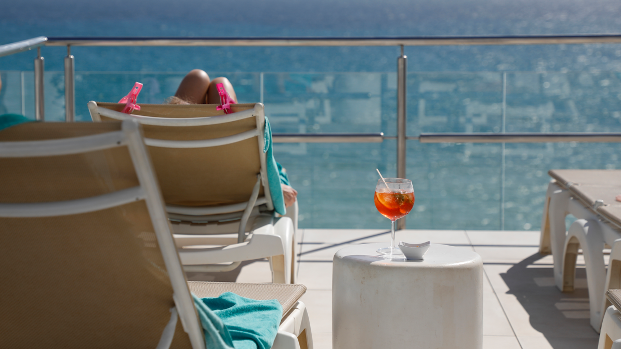 Eine Frau sonnt sich auf einer Liege. Neben ihr steht ein Aperol Spritz auf einem kleinen Tisch.