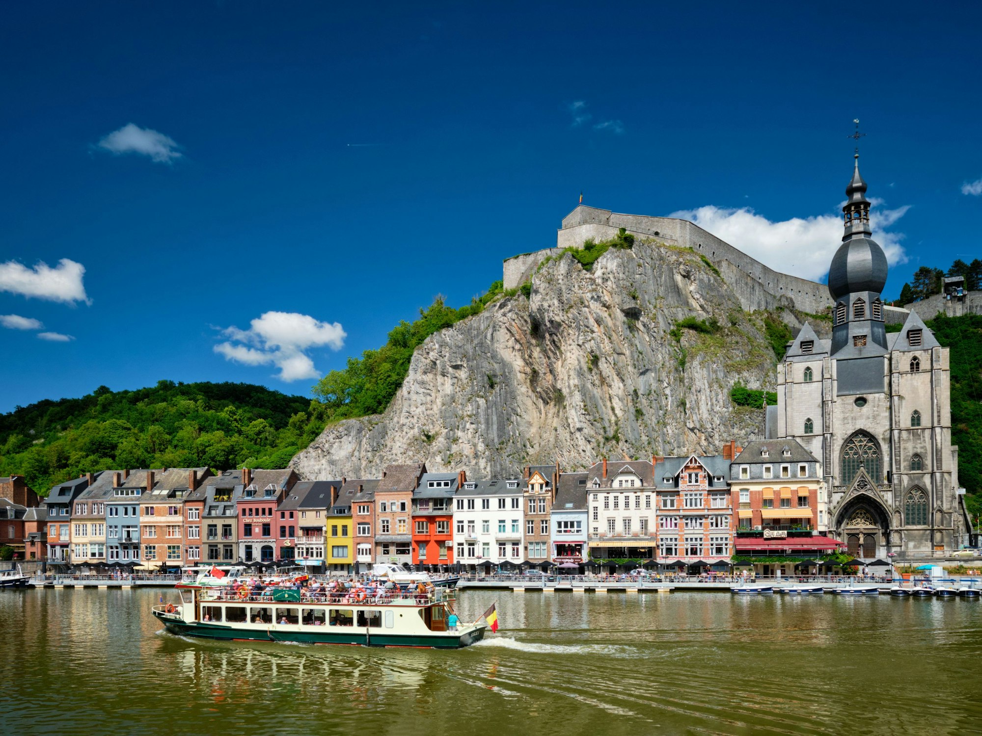 Blick über den Fluss auf die kunterbunten Gebäude der belgischen Stadt Dinant. Im Vordergrund fährt ein Boot mit Touristinnen und Touristen.