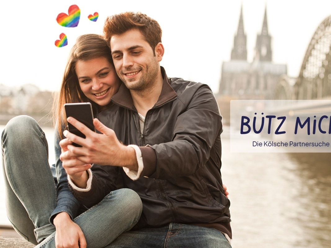 Eine Frau und ein Mann stehen auf der Rheinpromenade mit dem Kölner Dom und der Hohenzollernbrücke im Hintergrund und schauen lächelnd auf ein Smartphone. Daneben das Logo der Website Bütz mich.