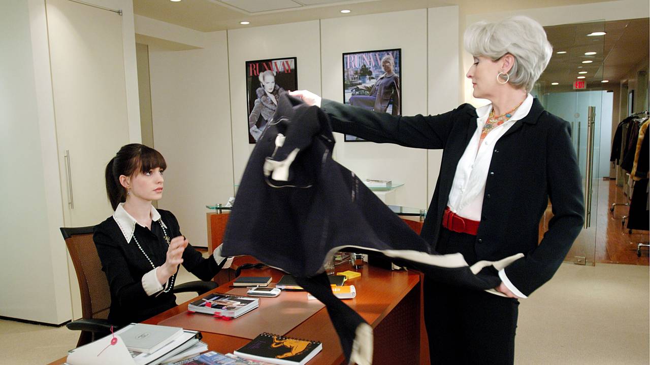 Miranda Priestly (Meryl Streep, r) wirft im Kinofilm „Der Teufel trägt Prada“ ihrer Assistentin Andrea Sachs (Anne Hathaway) die Jacke auf den Schreibtisch