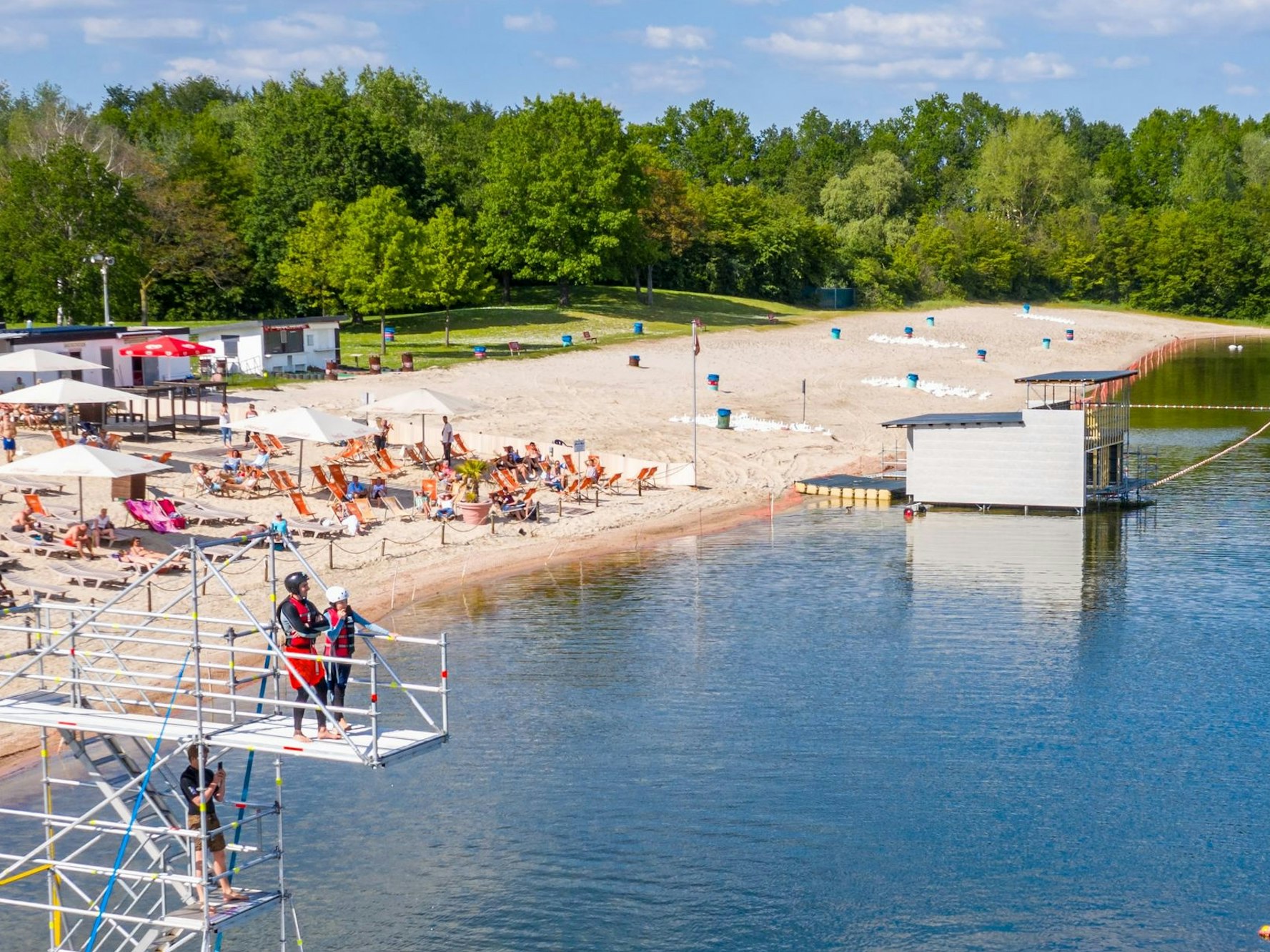 Ein Badesee mit Strand und Sprungturm, auf dem zwei Menschen stehen