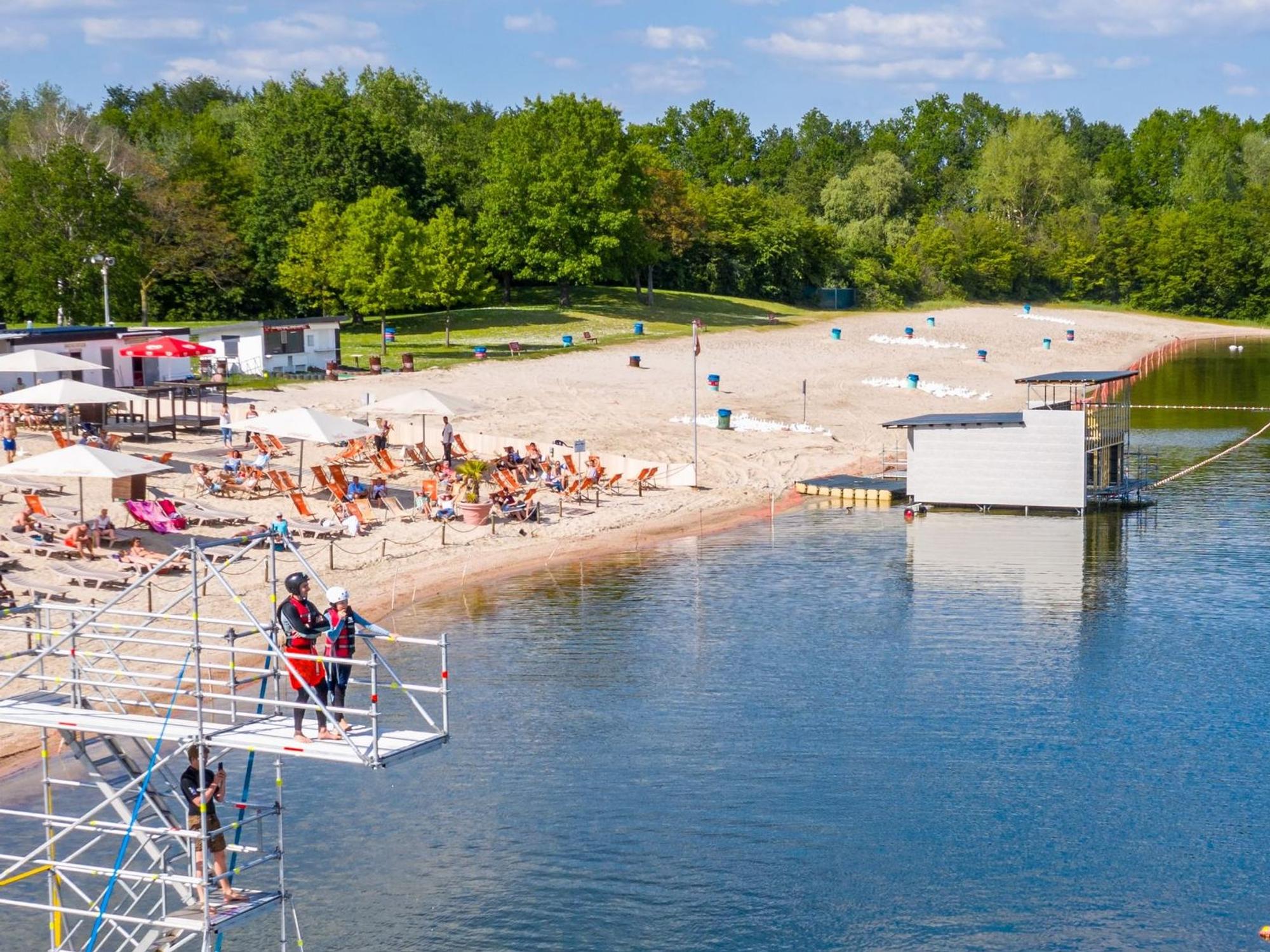Ein Badesee mit Strand und Sprungturm, auf dem zwei Menschen stehen