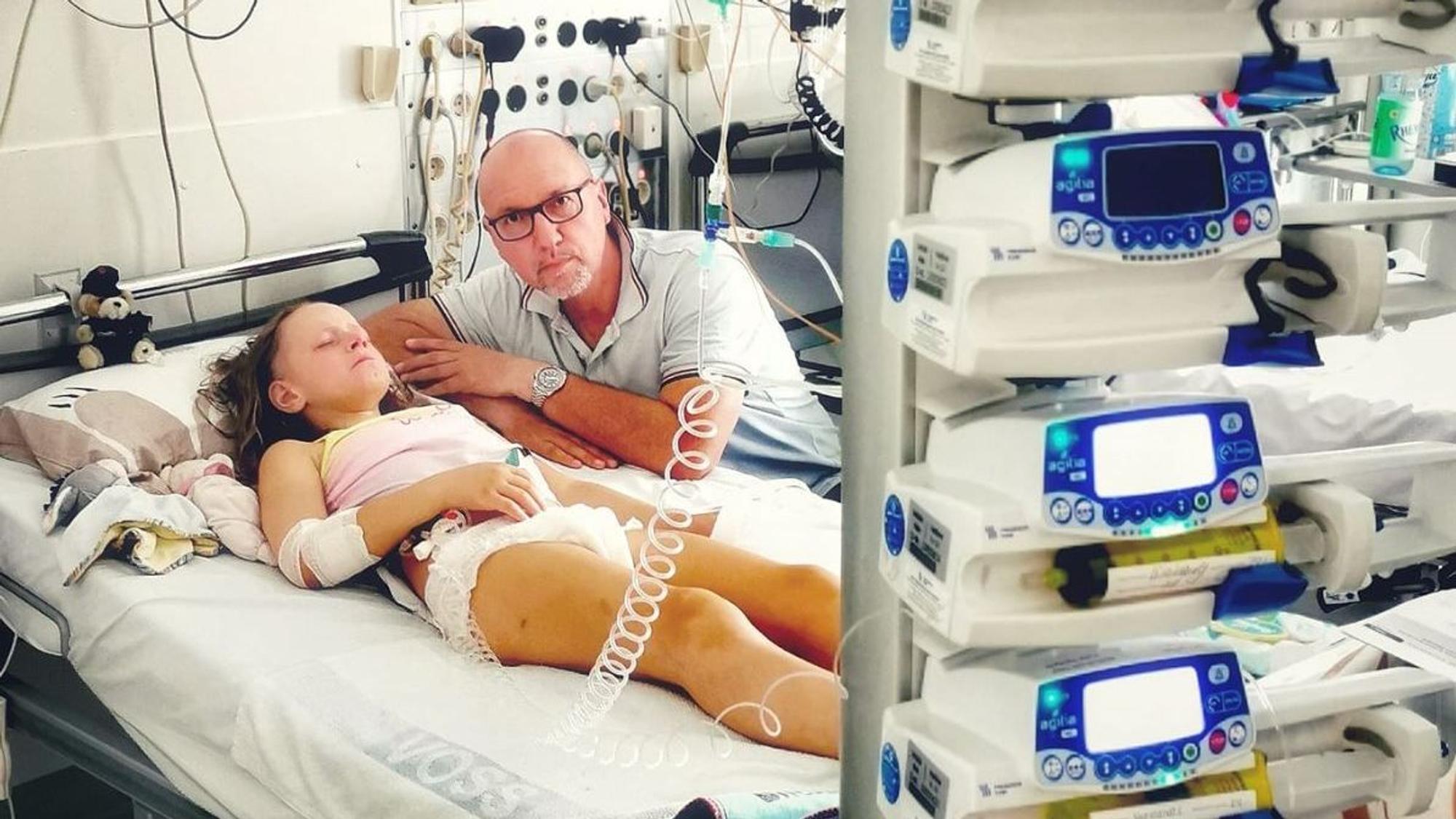 Lara aus Köln liegt an Geräten auf einem Krankenbett und schläft. Neben ihr schaut ein Mann in die Kamera.