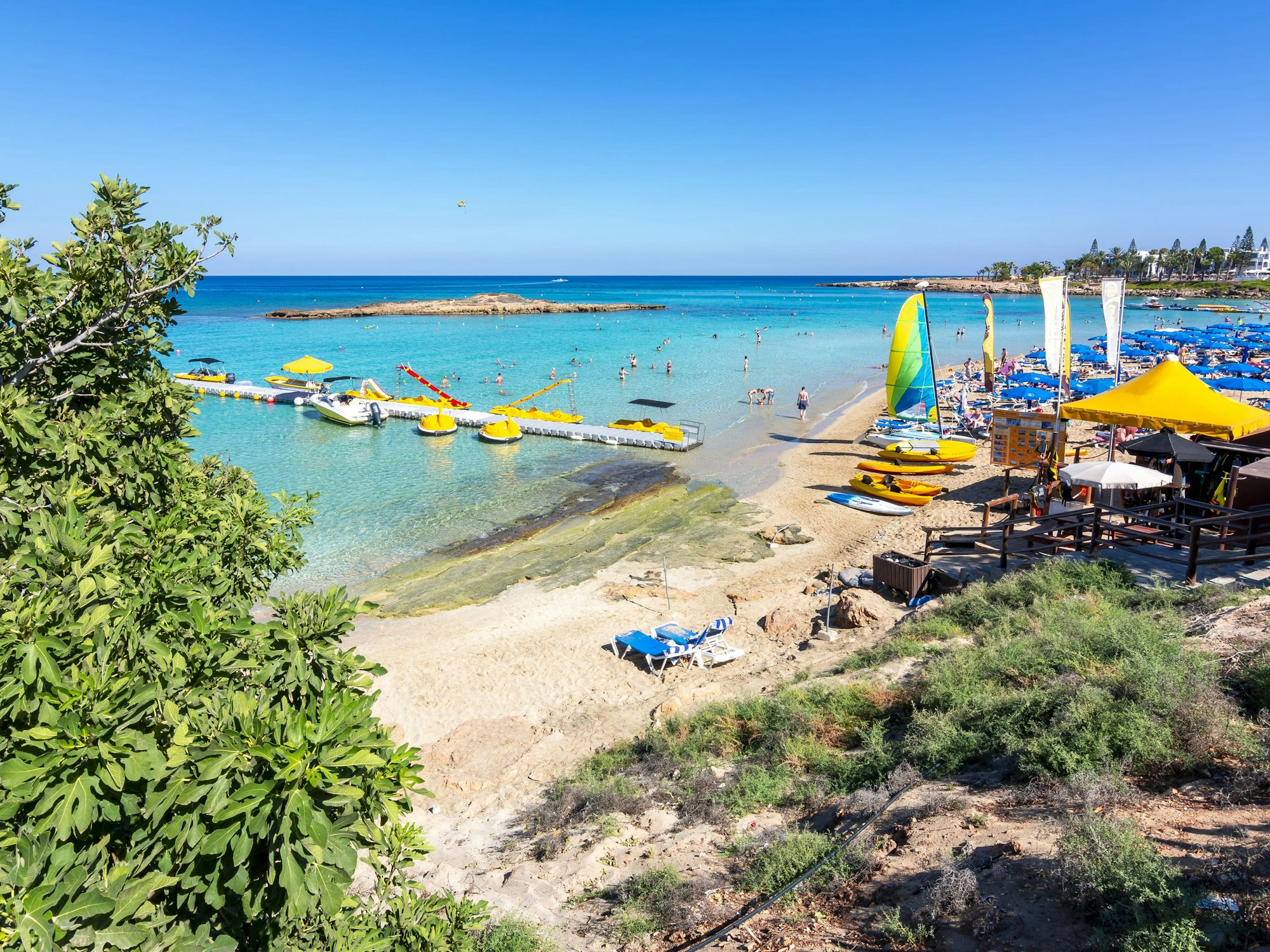 Sonnenliegen sowie Urlauberinnen und Urlauber in der Fig Tree Bay (Feigenbaumbucht) von Protaras auf Zypern. Die Fig Tree Bay (Feigenbaumbucht) von Protaras auf Zypern zählt zu den besten preiswerten Stränden in Europa.