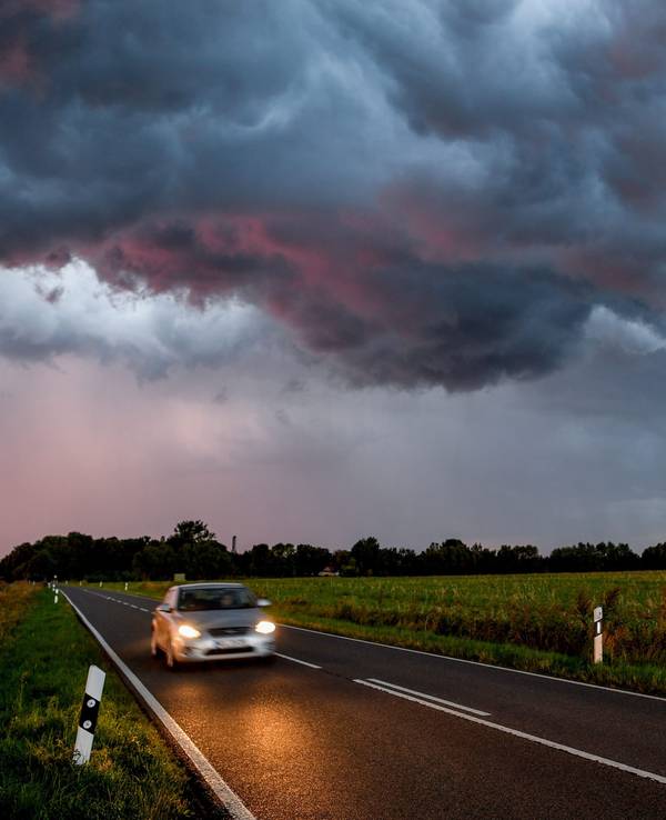 Ein Auto fährt auf einer Straße. Im Hintergrund nähert sich ein Unwetter mit Blitz und Donner.