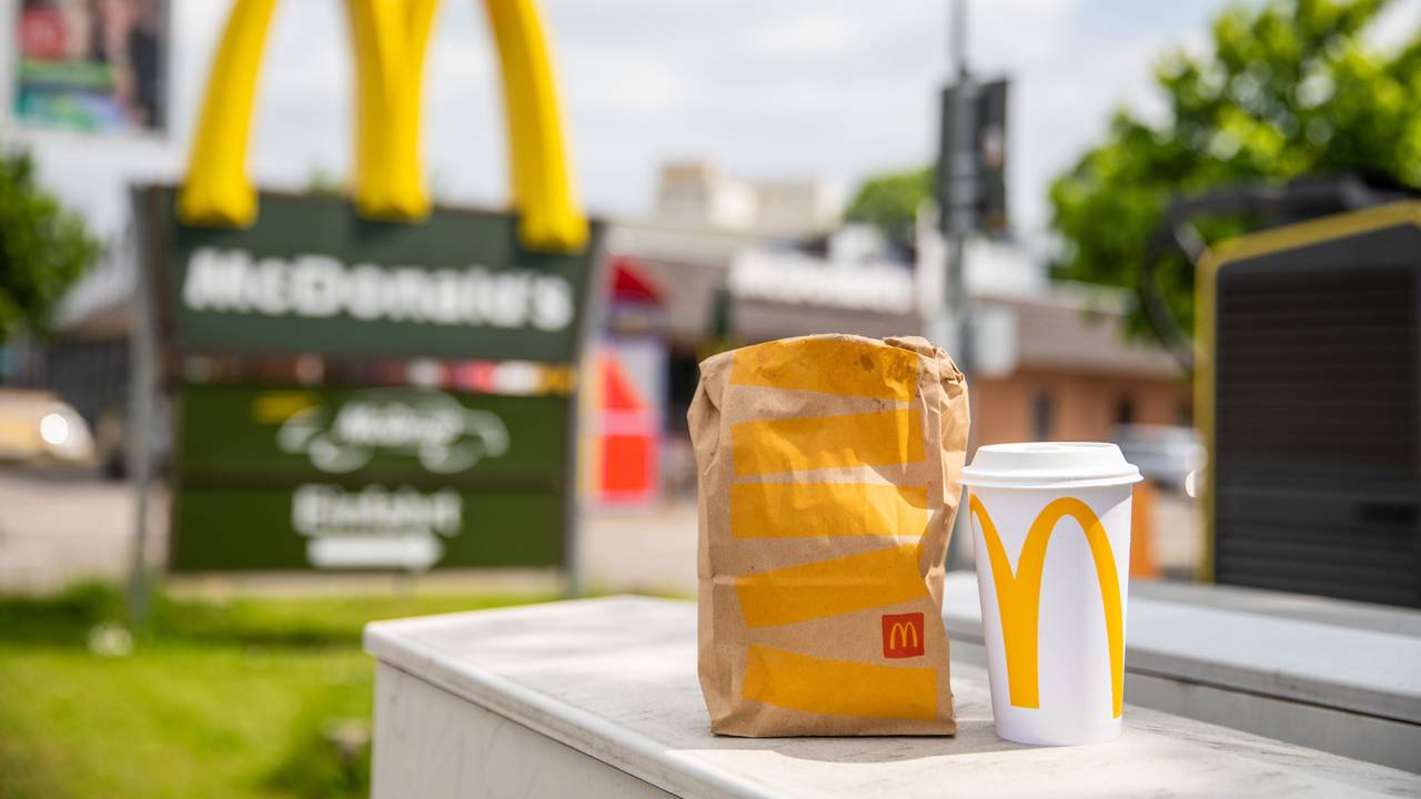 Eine McDonald’s To-Go-Tüte und ein dazugehöriger To-Go-Becher stehen vor einer McDonald’s-Filiale.