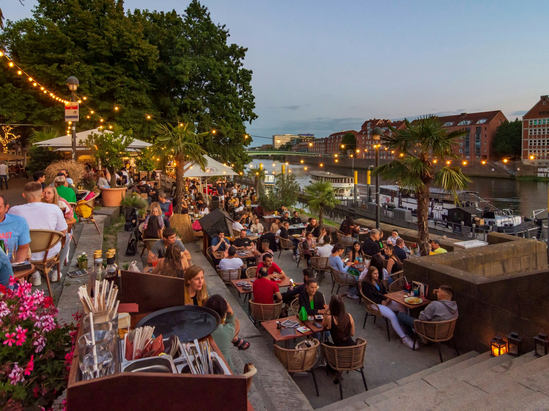 Eine mit Lichterketten und Palmen dekorierte Restaurantterrasse an der Weserpromenade in Bremen.