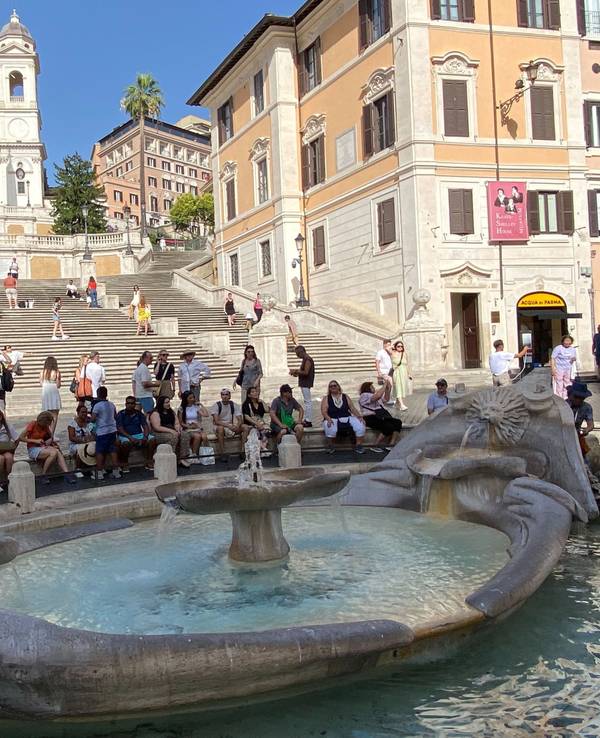 Touristinnen und Touristen sitzen im Schatten an der Fontana della Barcaccia, ein Brunnen auf der Piazza di Spagna, vor der Spanischen Treppe.