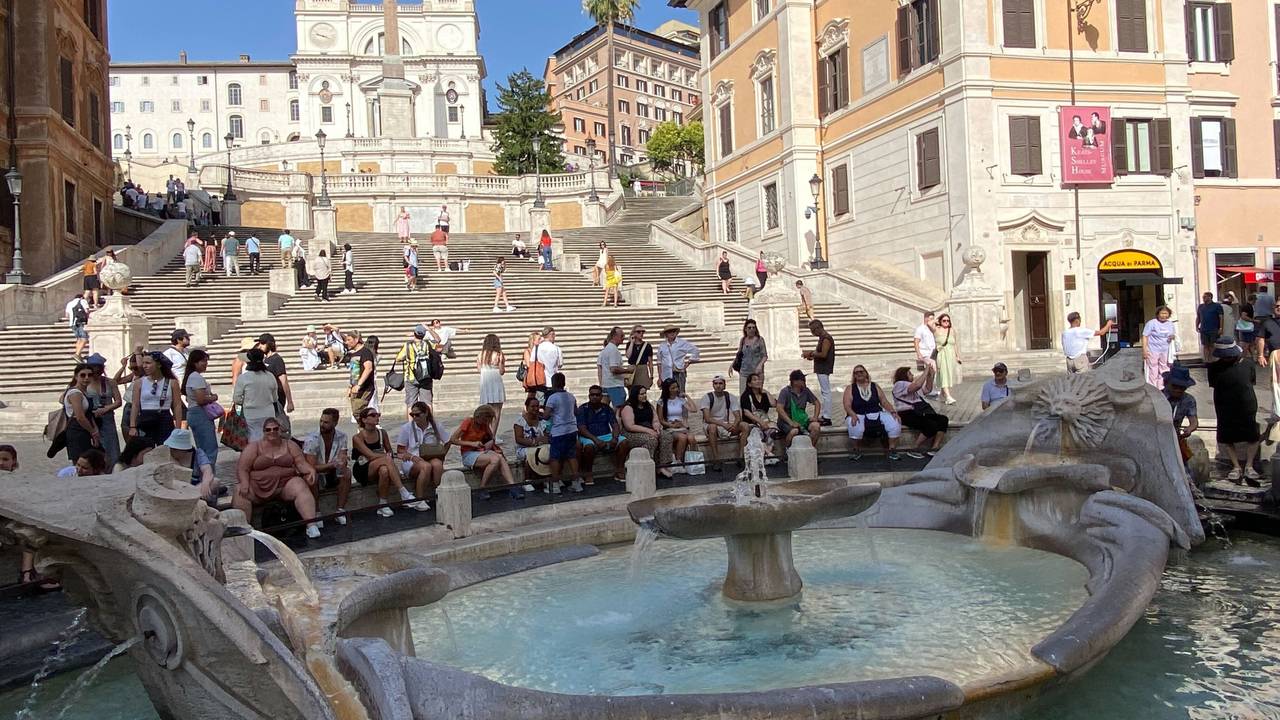 Touristinnen und Touristen sitzen im Schatten an der Fontana della Barcaccia, ein Brunnen auf der Piazza di Spagna, vor der Spanischen Treppe.