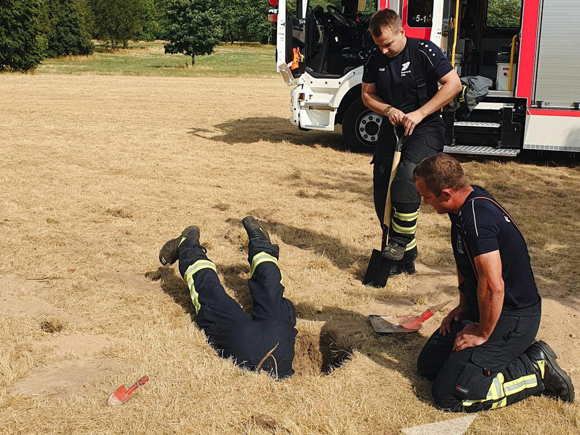 Ein Feuerwehrmann steckt kopfüber in einem Loch, ein Kollege kniet daneben, ein zweiter steht und hat eine Schaufel in der Hand.
