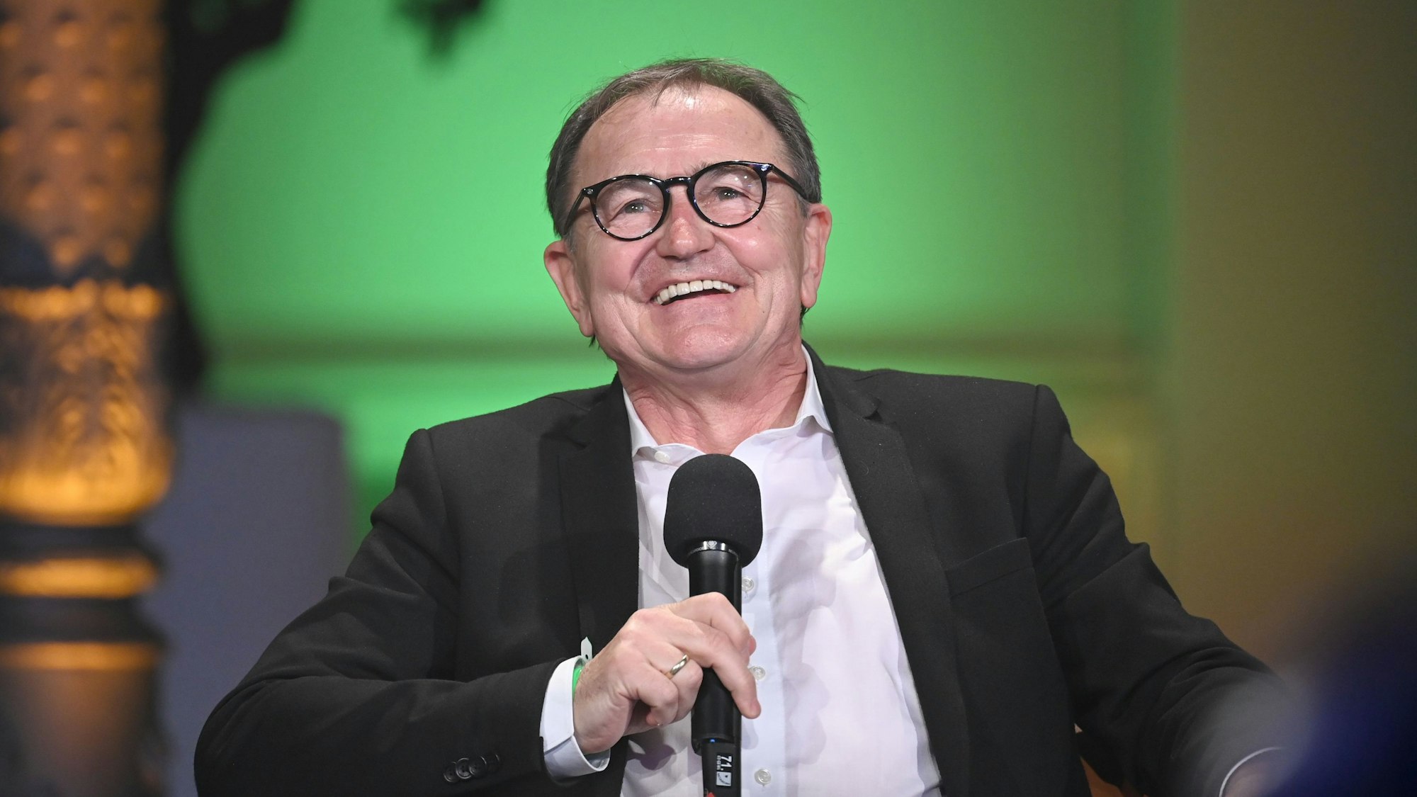 Ewald Lienen sitzt auf einer Bühne und hält ein Mikrofon in der Hand.