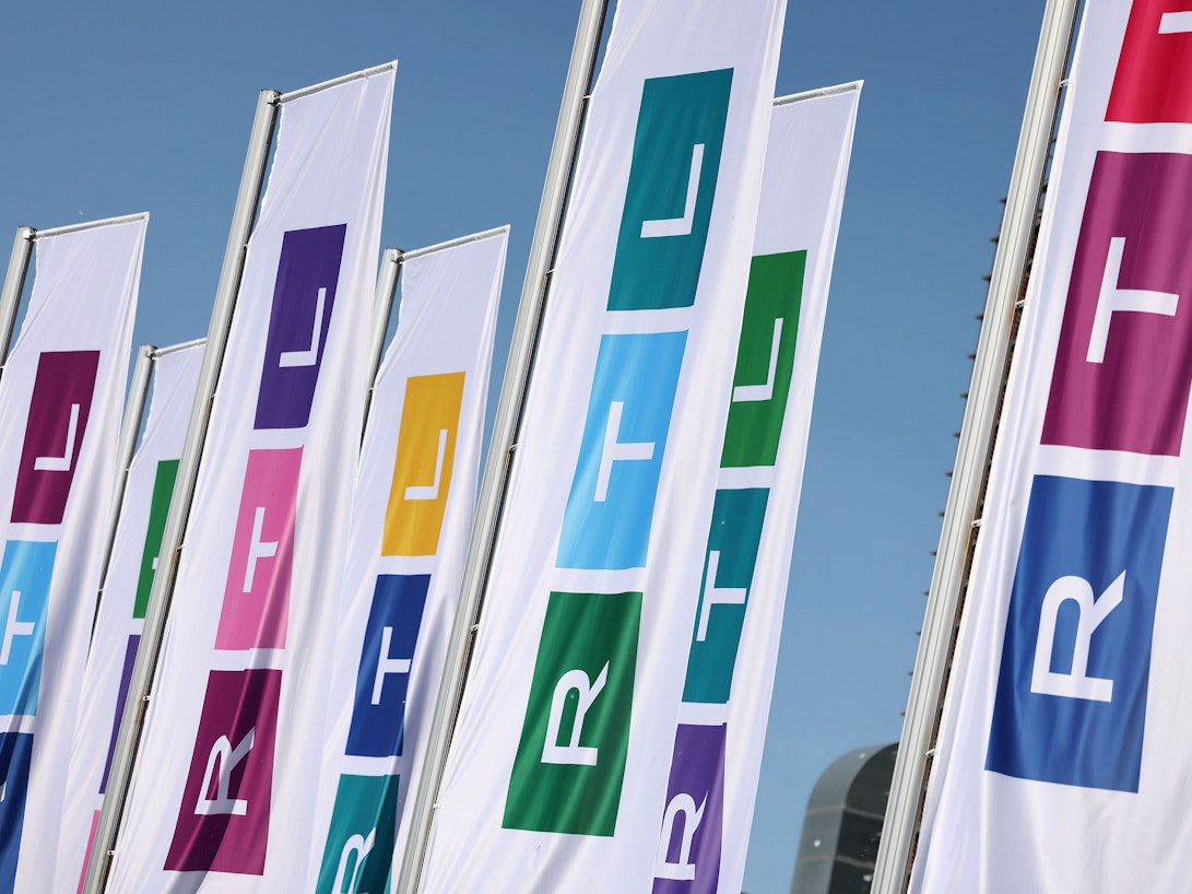 Fahnen mit dem neuen Logo des Fernsehsenders RTL wehen vor dem Gebäude. Die drei Buchstaben des Privatsenders sind schmaler, statt aus den drei Grundfarben Rot, Gelb, Blau sind die drei Blöcke des Logos aus allen möglichen Farben zusammensetzen.