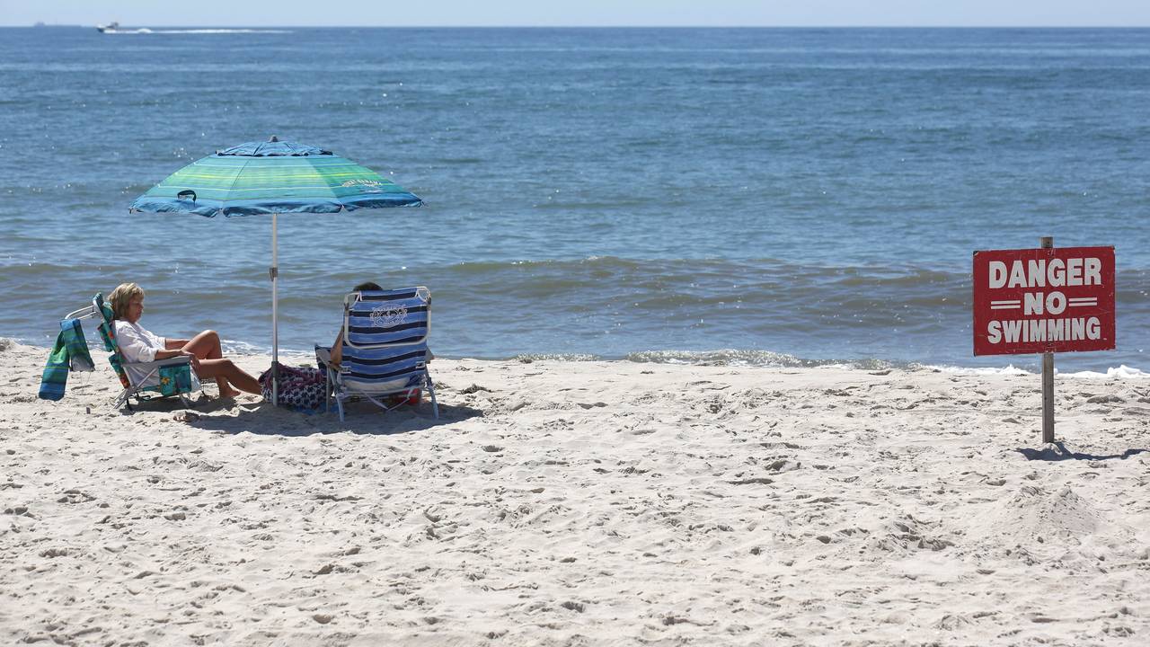 Zwei Menschen sitzen am Strand, neben ihnen steht ein Schild mit der Aufschrift "Vorsicht, nicht schwimmen".