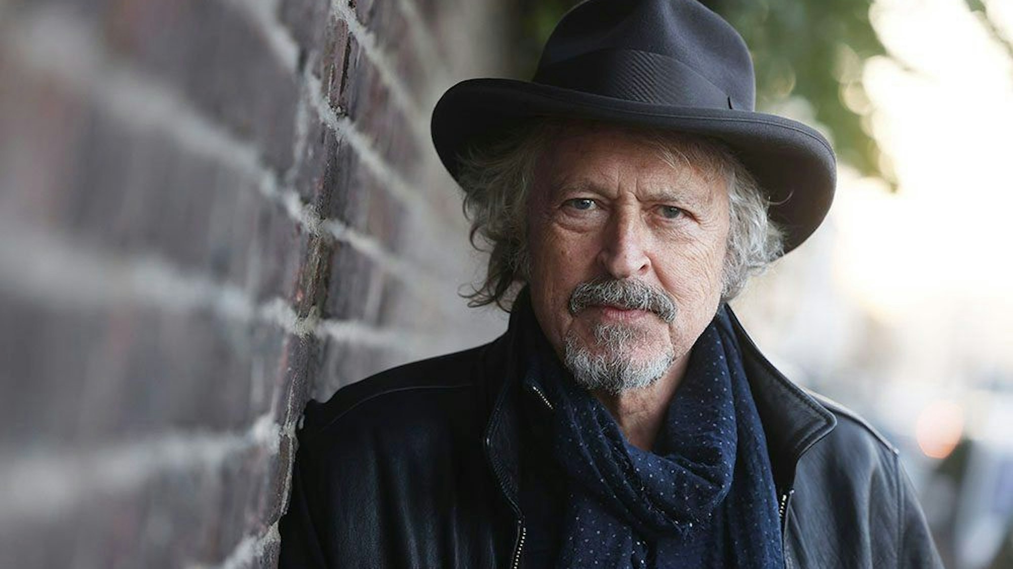 Wolfgang Niedecken lehnt mit Hut an einer Steinmauer und schaut in die Kamera.