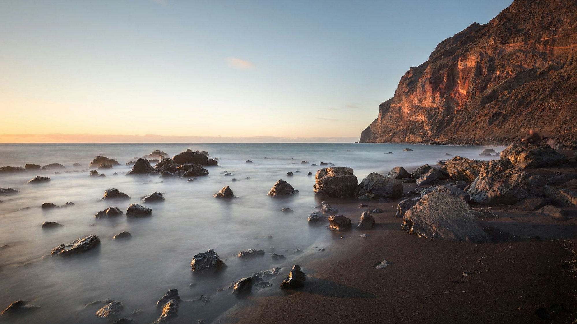 Ein dunkler Sandstrand mit einigen Felsen. Im Hintergrund hohe Klippen.
