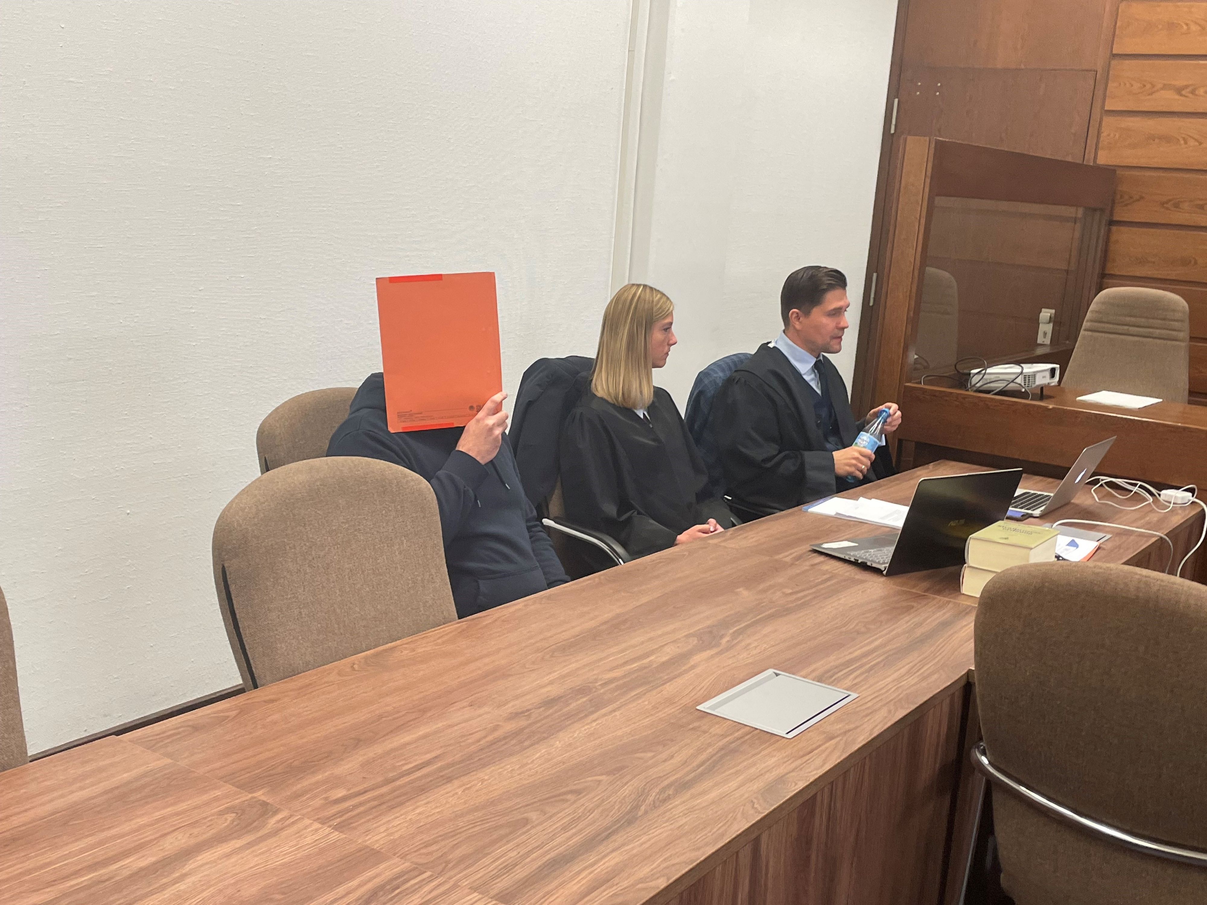 Der Angeklagte hält sich einen Heftordner vor das Gesicht, neben ihm sitzen seine Anwältin und sein Anwalt.