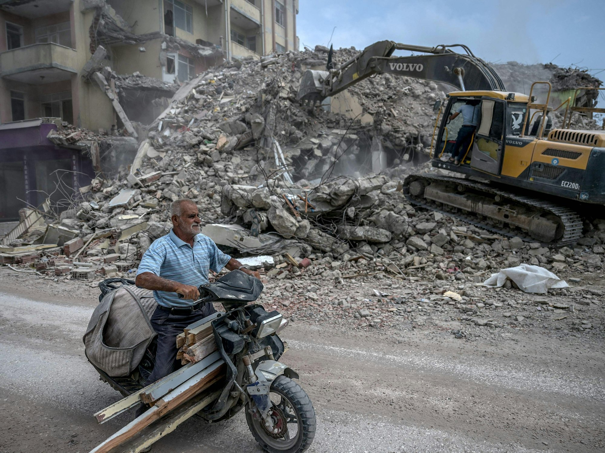 Noch immer liegt vielerorts meterhoher Schutt in den Straßen, es sieht so aus, als wäre das Erdbeben nur Wochen her – und nicht Monate. Auf dem Foto fährt ein Mann am 9. Juli durch die Straßen von Samandag im Südosten der Türkei, während ein Bagger sich durch Schutt gräbt.
