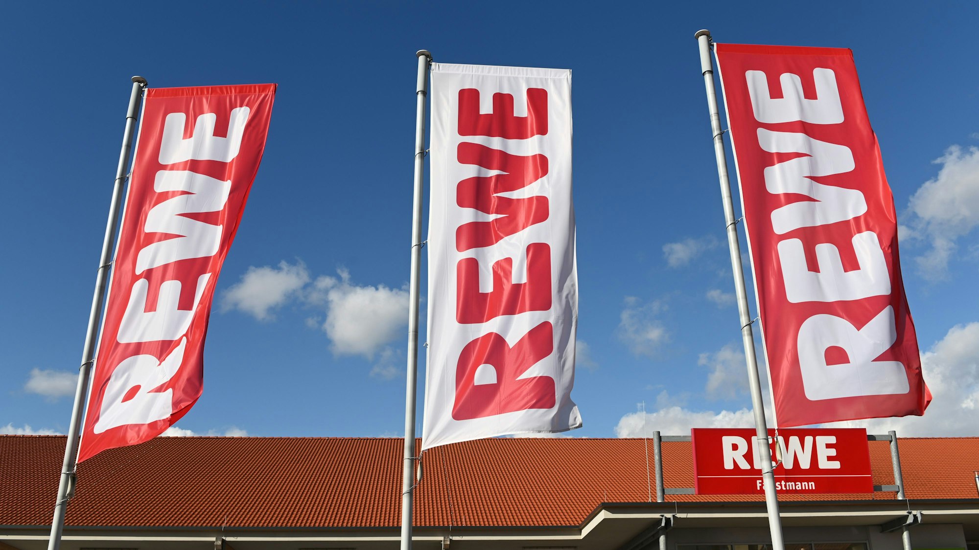 Fahnen mit dem Logo des Lebensmitteleinzelhändlers Rewe wehen vor einer Filiale.