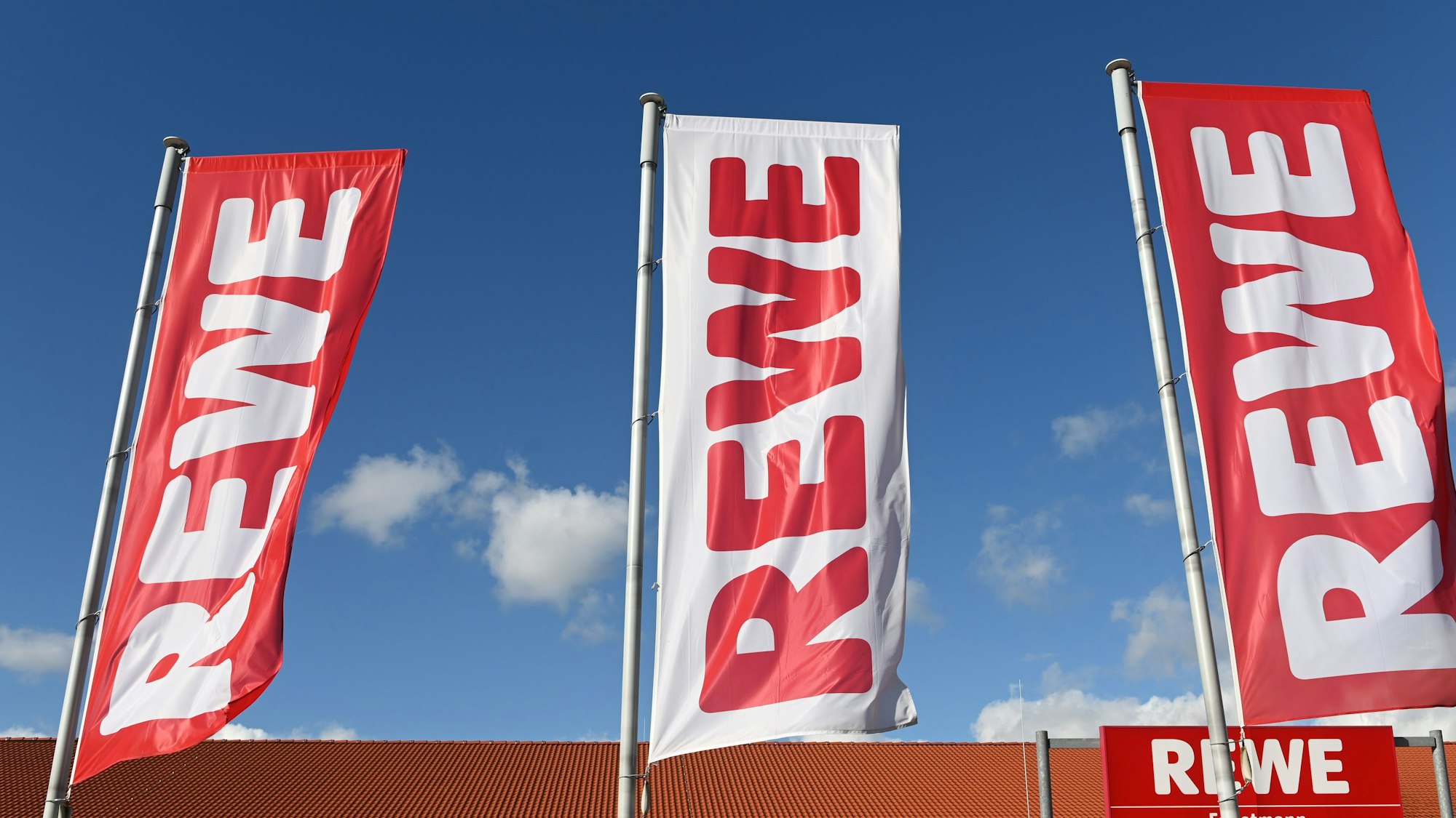Fahnen mit dem Logo des Lebensmitteleinzelhändlers Rewe wehen vor einer Filiale.