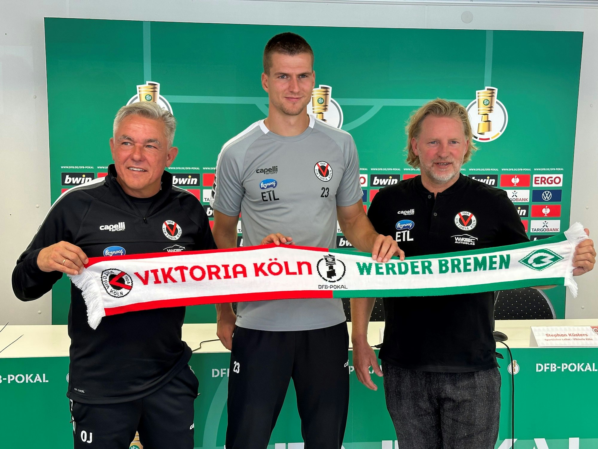 Viktoria-Trainer Olaf Janßen, Kapitän Moritz Fritz und Sportdirektor Stephan Küsters halten den Spieltagsschal in die Kamera.