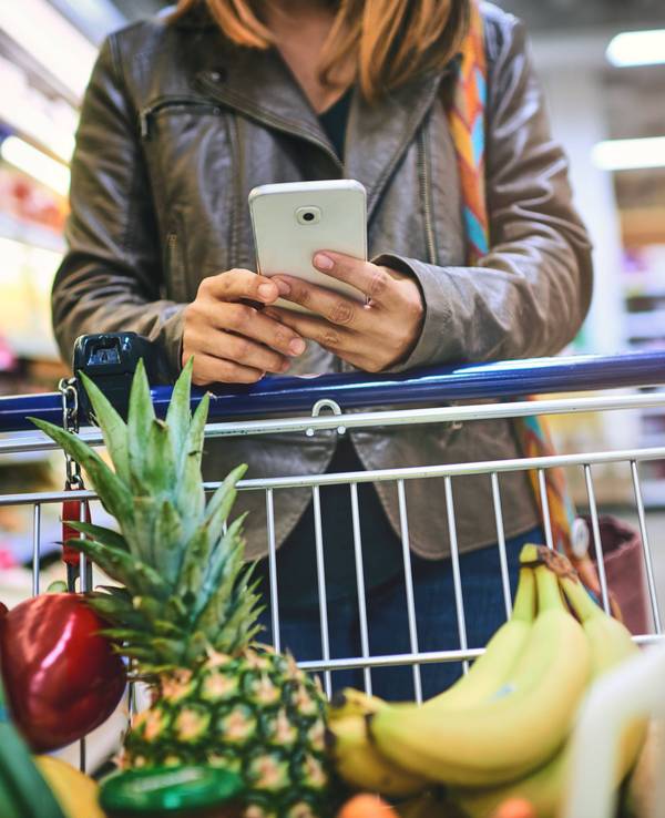 Eine Frau benutzt ihr Smartphone während sie sich auf einem gefüllten Einkaufswagen abstützt.