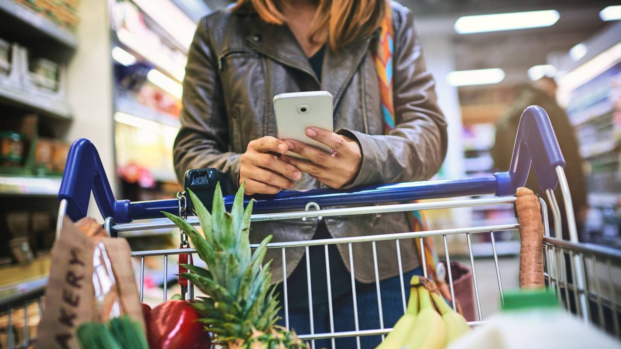 Eine Frau benutzt ihr Smartphone während sie sich auf einem gefüllten Einkaufswagen abstützt.