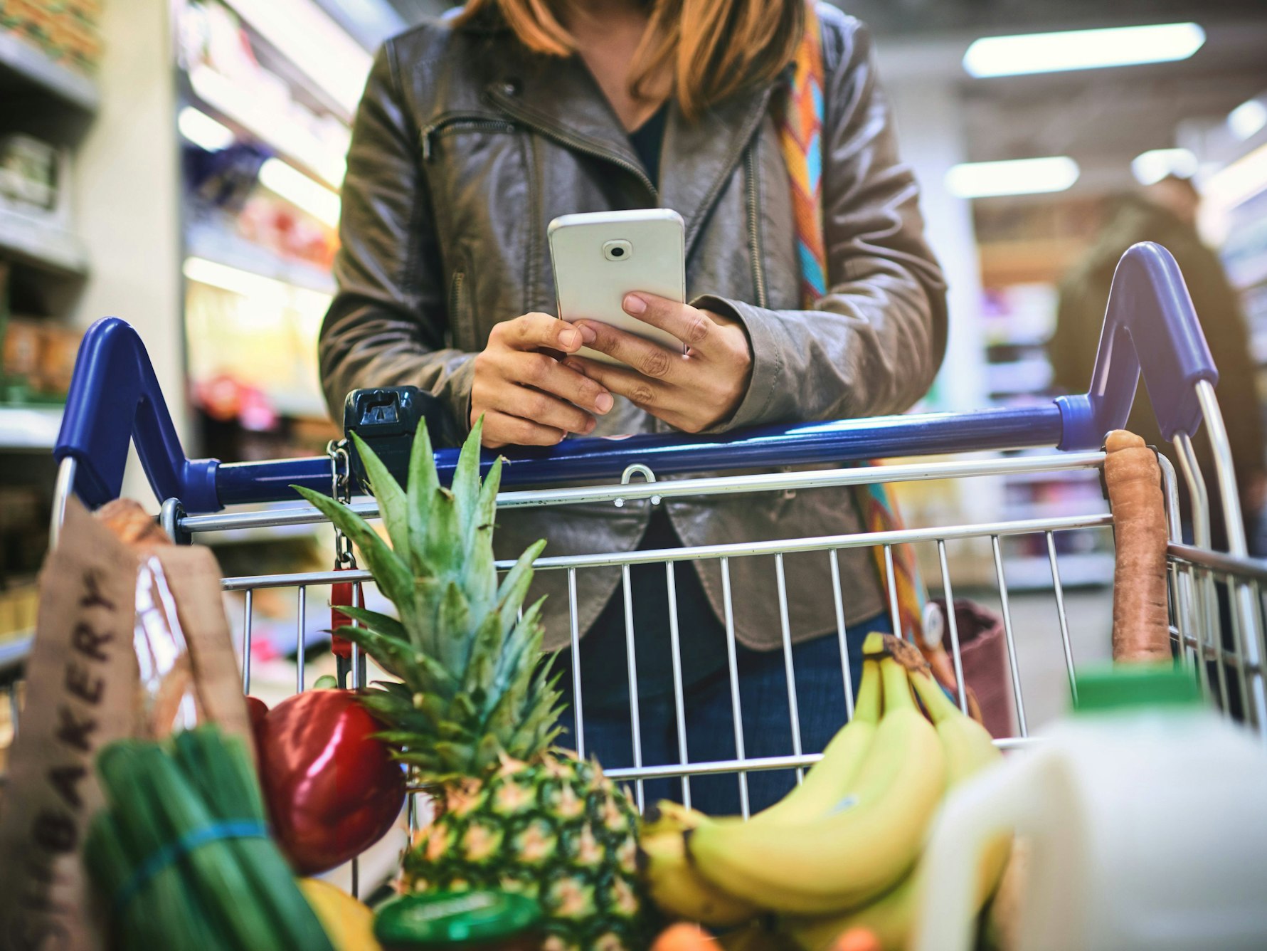 Eine Frau lehnt mit ihrem Smartphone auf einem gefüllten Einkaufswagen.