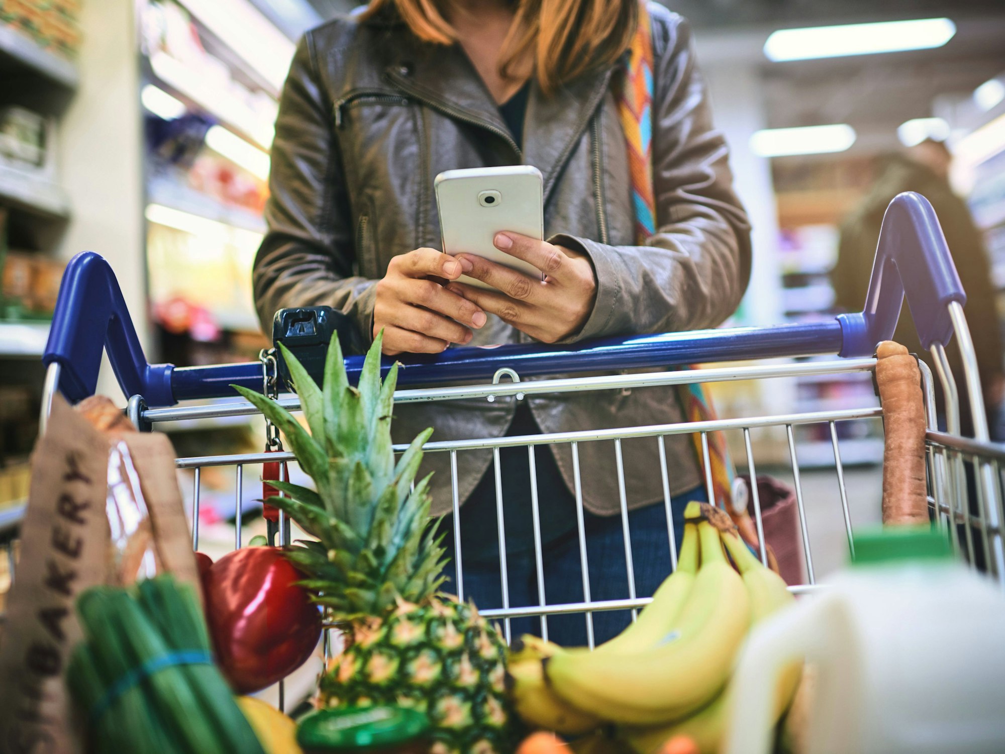 Eine Frau benutzt ihr Smartphone während sie sich auf einem gefüllten Einkaufswagen abstützt.