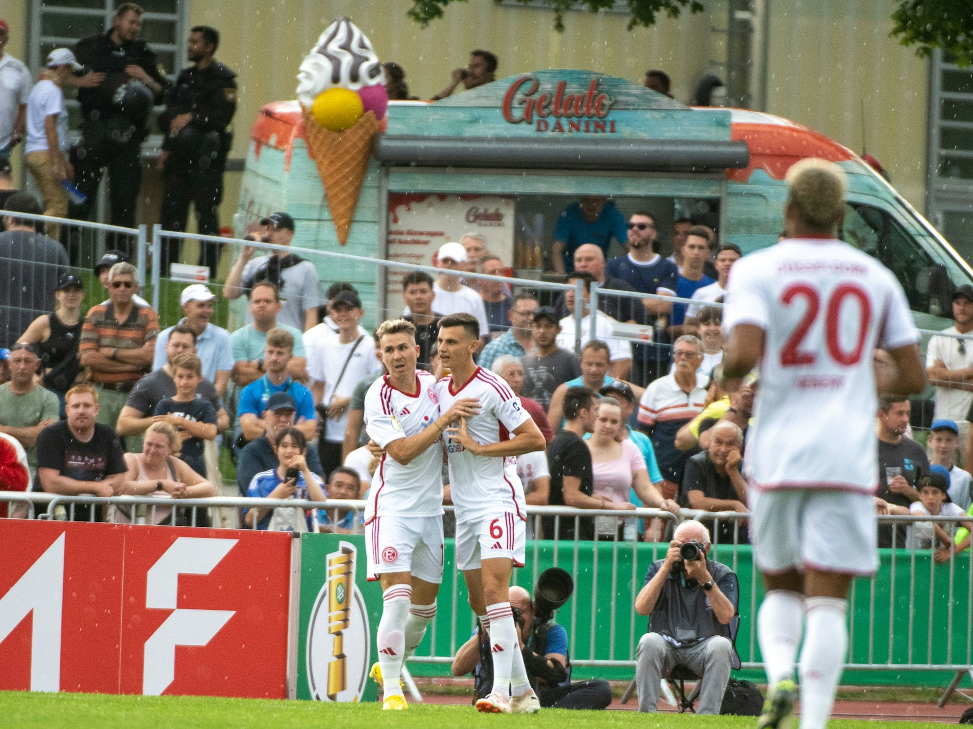 Düsseldorfs Felix Klaus (l) und Yannik Engelhardt jubeln nach dem 1:0. Im Hintergrund stehen ein paar wenige Zuschauerinnen und Zuschauer sowie ein Eiswagen.