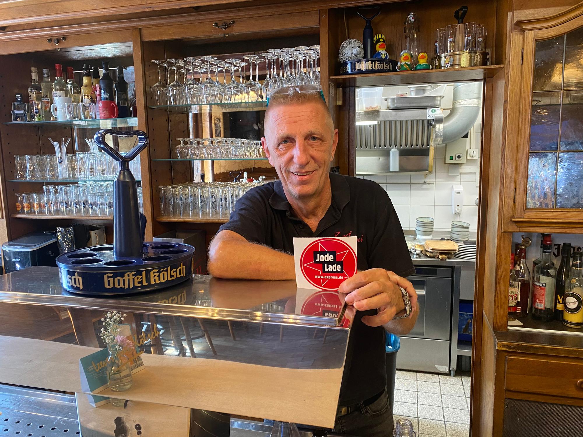 Ralf Klein steht hinter der Theke und zeigt die „Jode Lade“-Auszeichnung