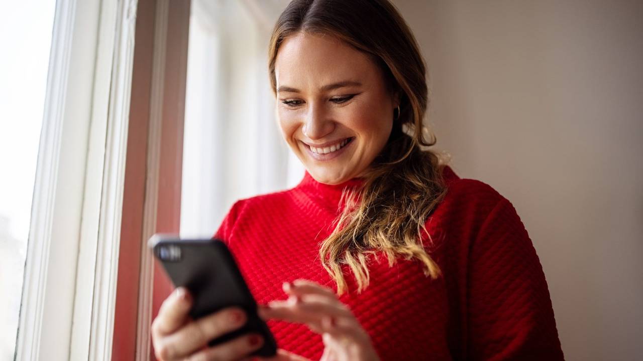 Frau trägt roten Pullover, hält Handy in der Hand und lächelt.