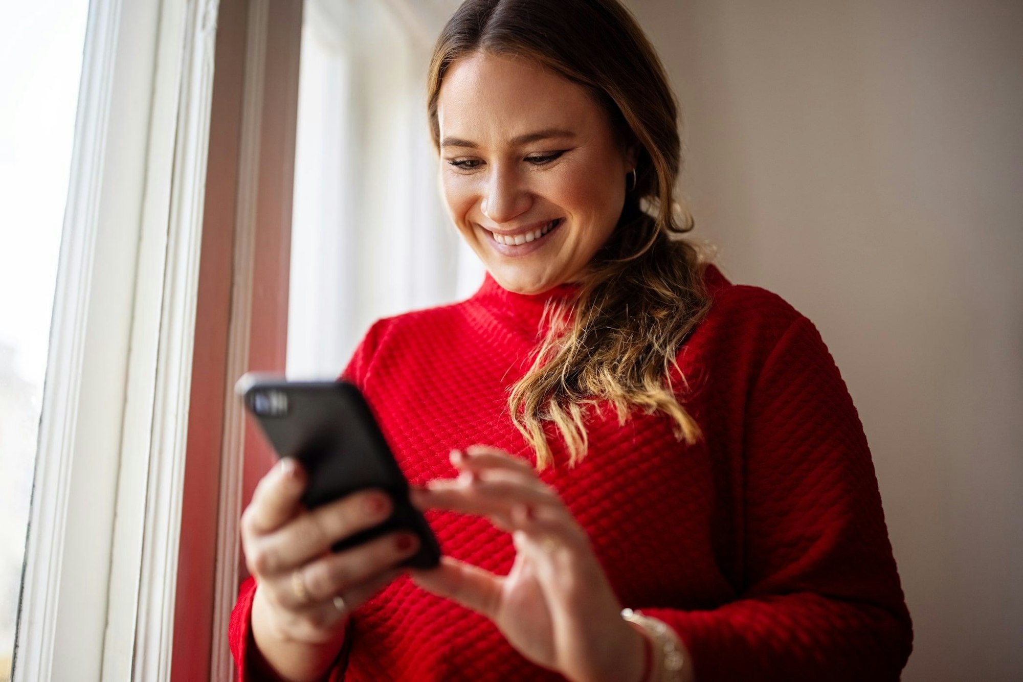 Frau trägt roten Pullover, hält Handy in der Hand und lächelt.