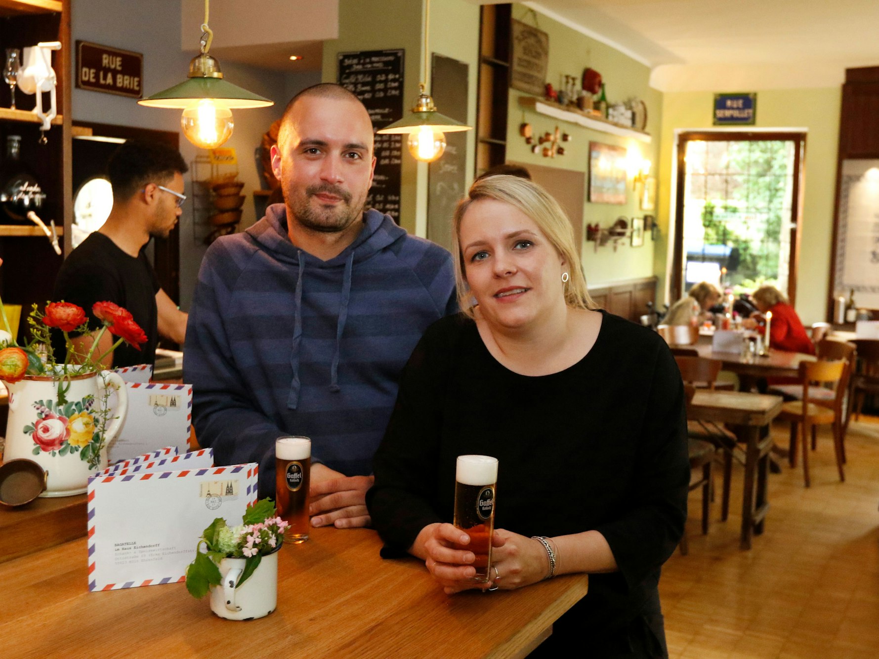 Reja und Daniel Rabe stehen mit Kölschgläsern an einem Tresen und lächeln in die Kamera.