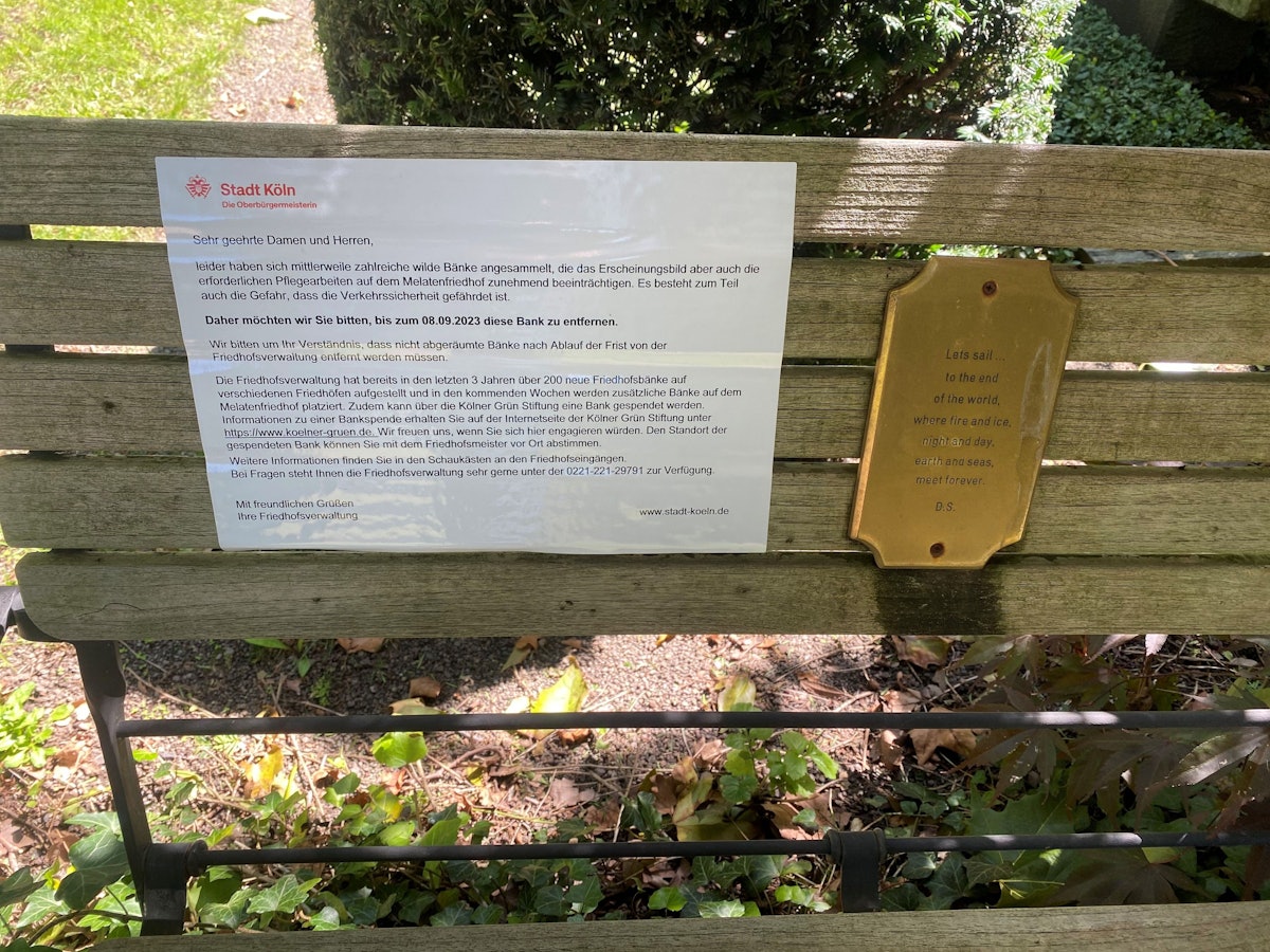 Eine Bank auf dem Melatenfriedhof. Auf ihr ist ein Hinweisschild der Stadt Köln angebracht, dass sie entfernt werden soll.