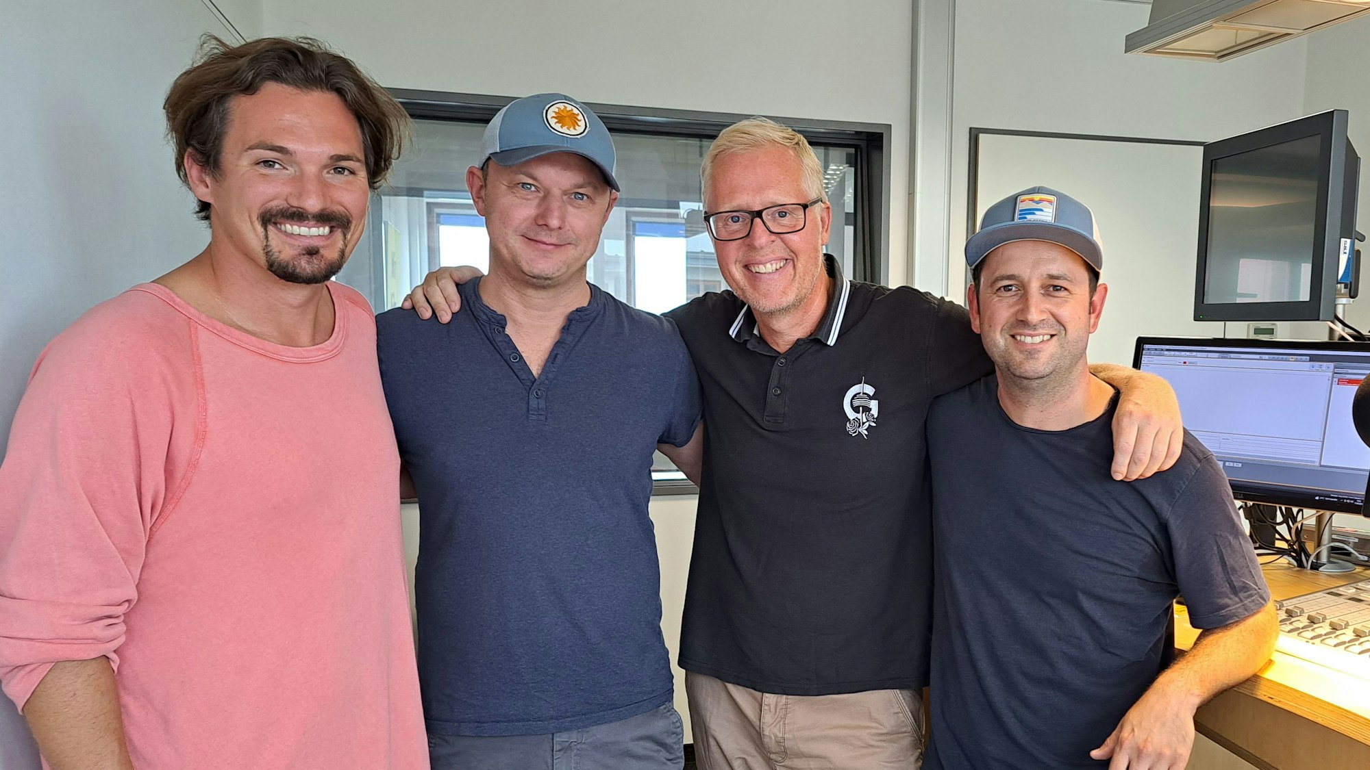 Vier Männer halten sich im Arm und lächeln in einem Tonstudio in die Kamera.