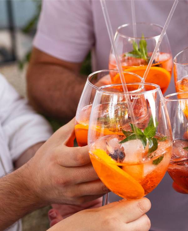 Vier Personen stoßen mit Gläsern, die mit dem orangen Aperol Spritz gefüllt sind, an.