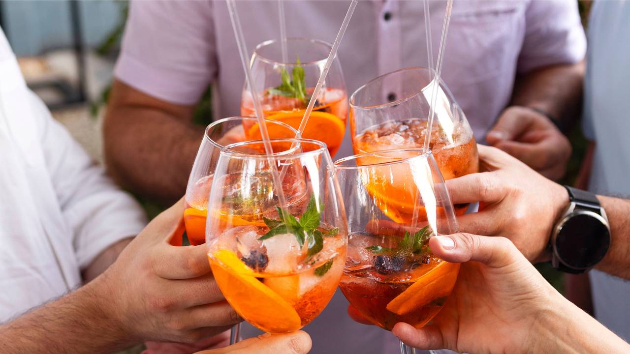 Vier Personen stoßen mit Gläsern, die mit dem orangen Aperol Spritz gefüllt sind, an.