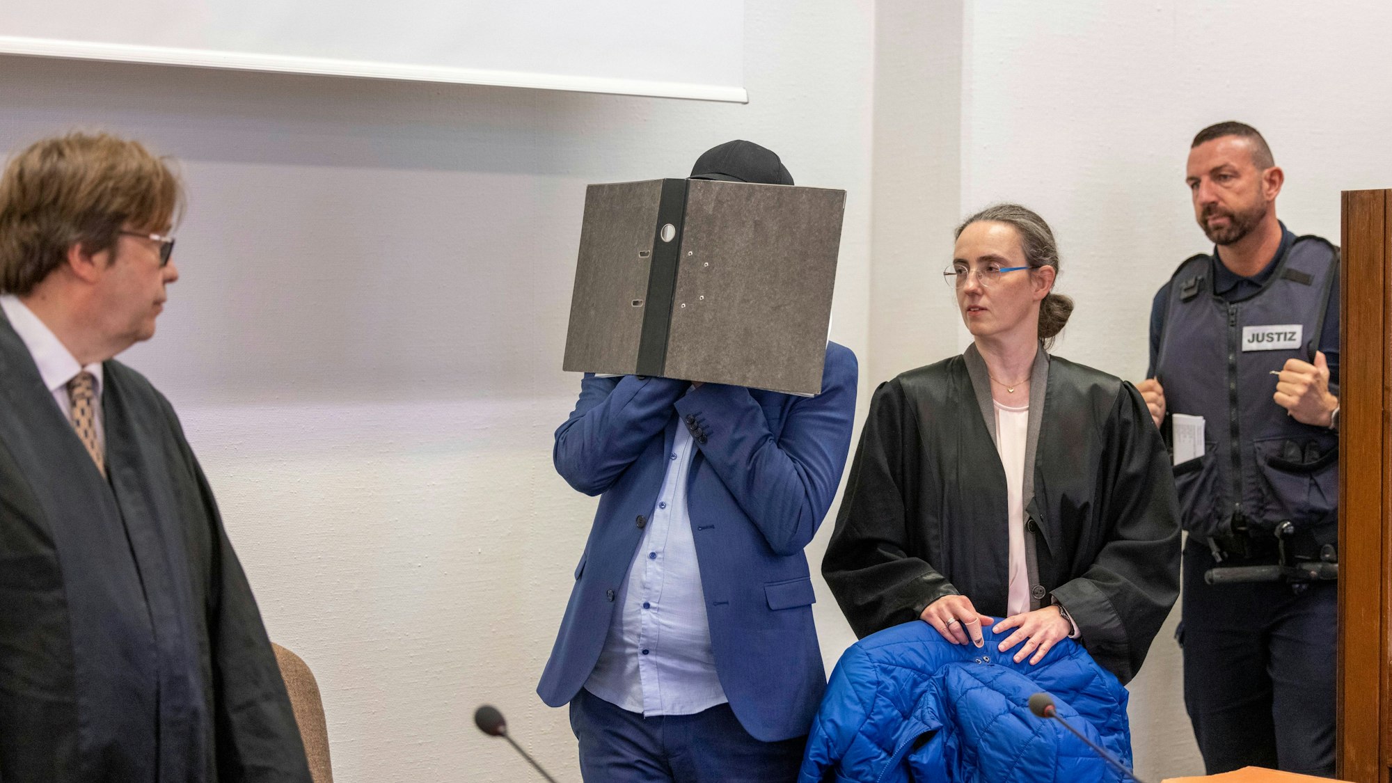 Angeklagter hält sich in Kölner Gericht einen Aktenordner vor das Gesicht. Daneben sind seine Anwälte und ein Polizist zu sehen.