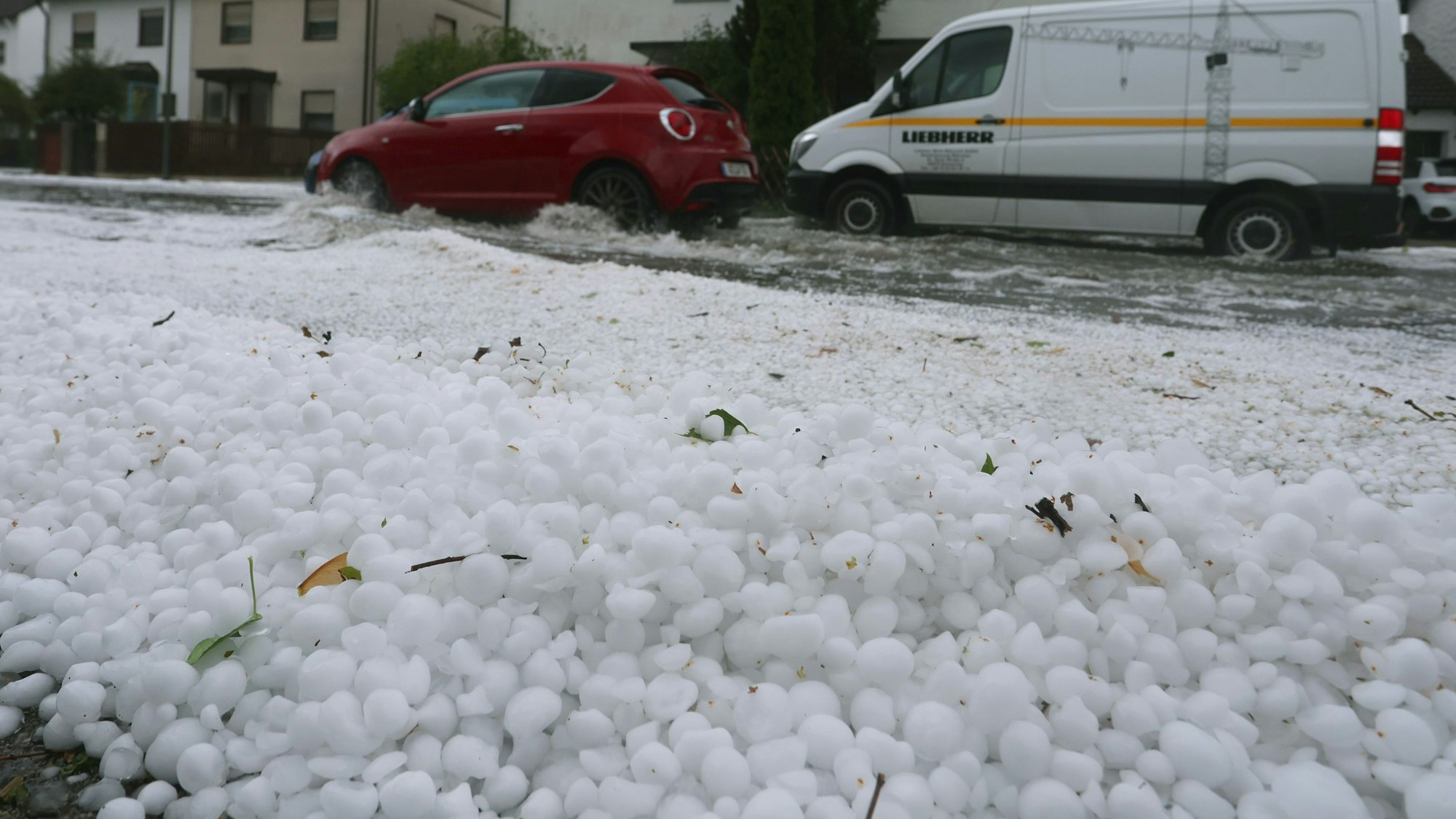 Hagel liegt auf einem Gehsteig und treibt auf einer überfluteten Straße.